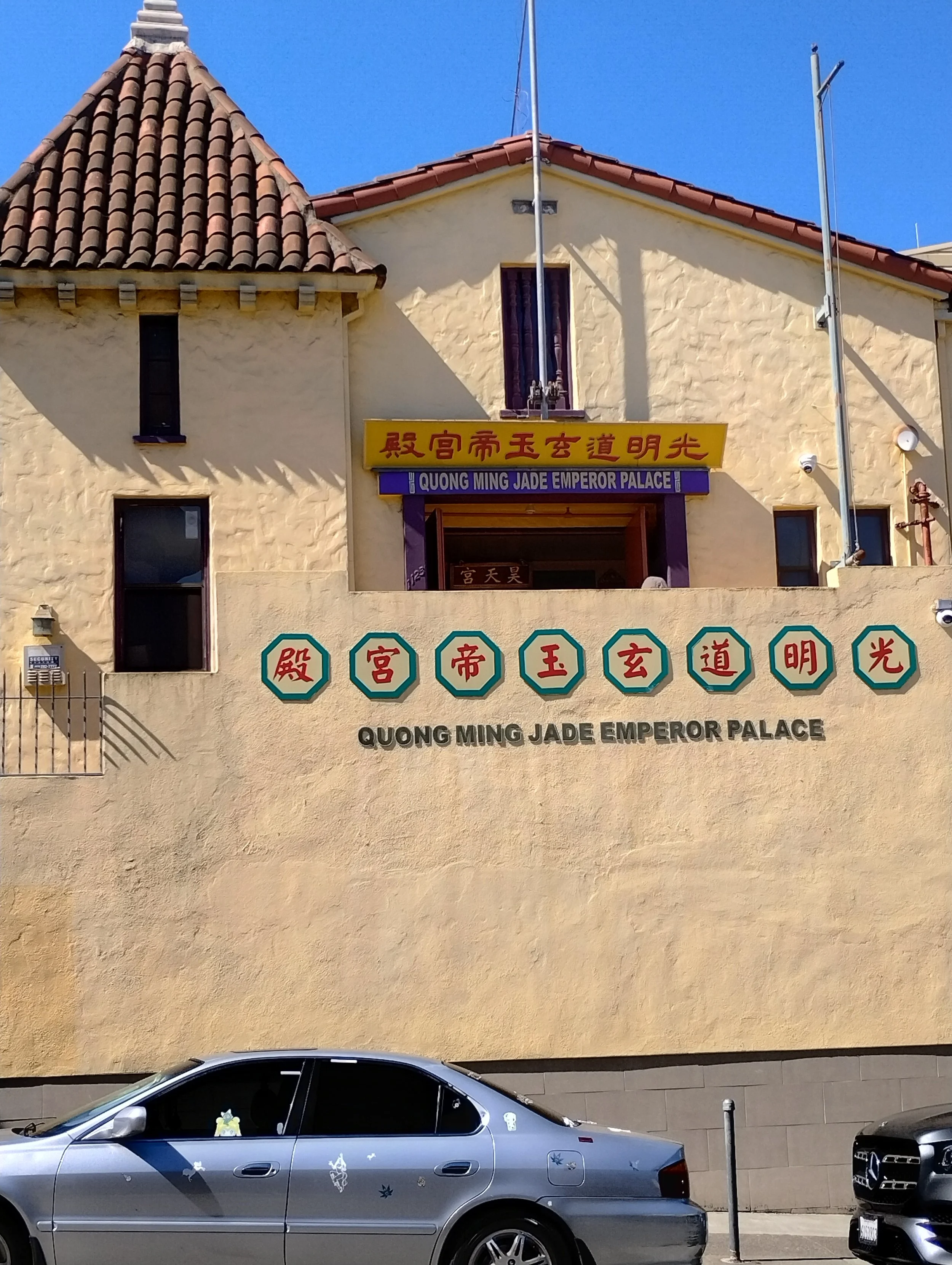 Exterior view of the Quong Ming Jade Emperor Palace building with a yellow sign and Chinese characters, and a parking lot with a gray car decorated with stickers in the foreground, under a clear blue sky.