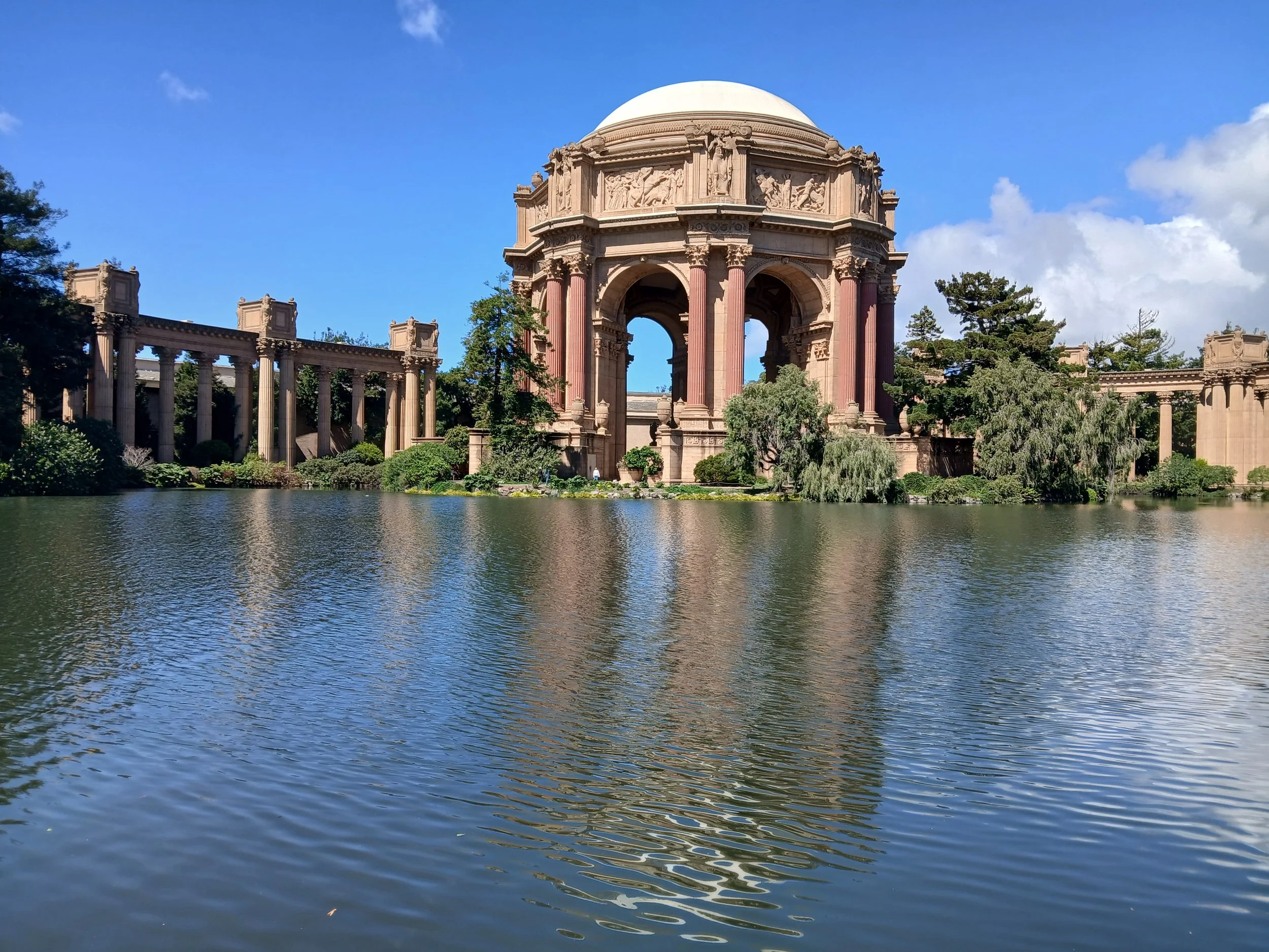 A historic temple with an ornate facade and a dome, surrounded by trees, situated by a body of water under a partly cloudy sky.