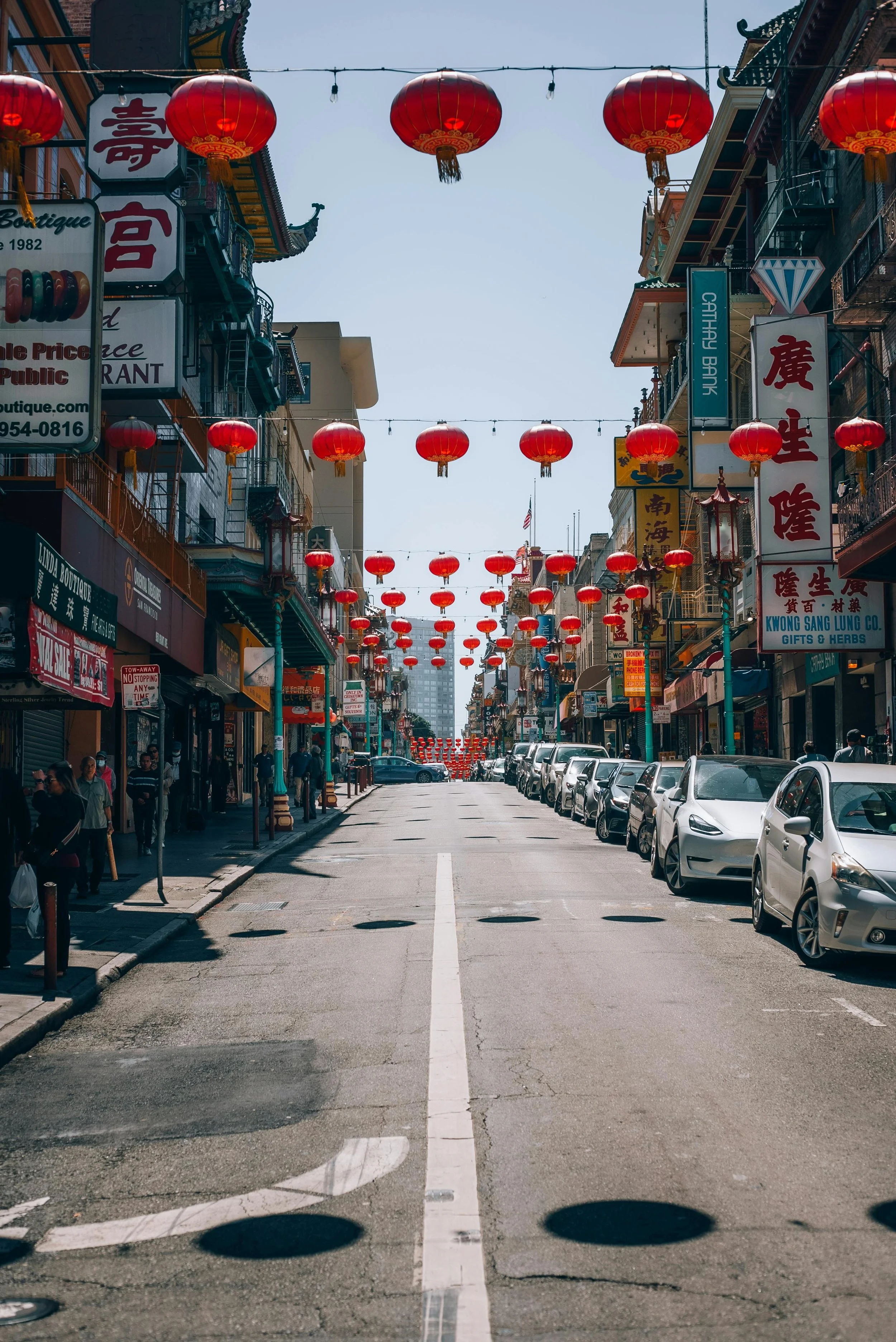 A city street decorated with red Chinese lanterns hanging above on strings. There are buildings on both sides with signs, some with Chinese characters, and parked cars along the curb. People are walking on the sidewalks.