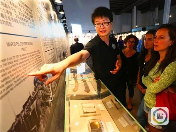 A man is explaining an exhibit at a museum or gallery to a group of women. The exhibit includes historical documents and artifacts displayed in glass cases. The man is pointing at a large wall display with typed text.