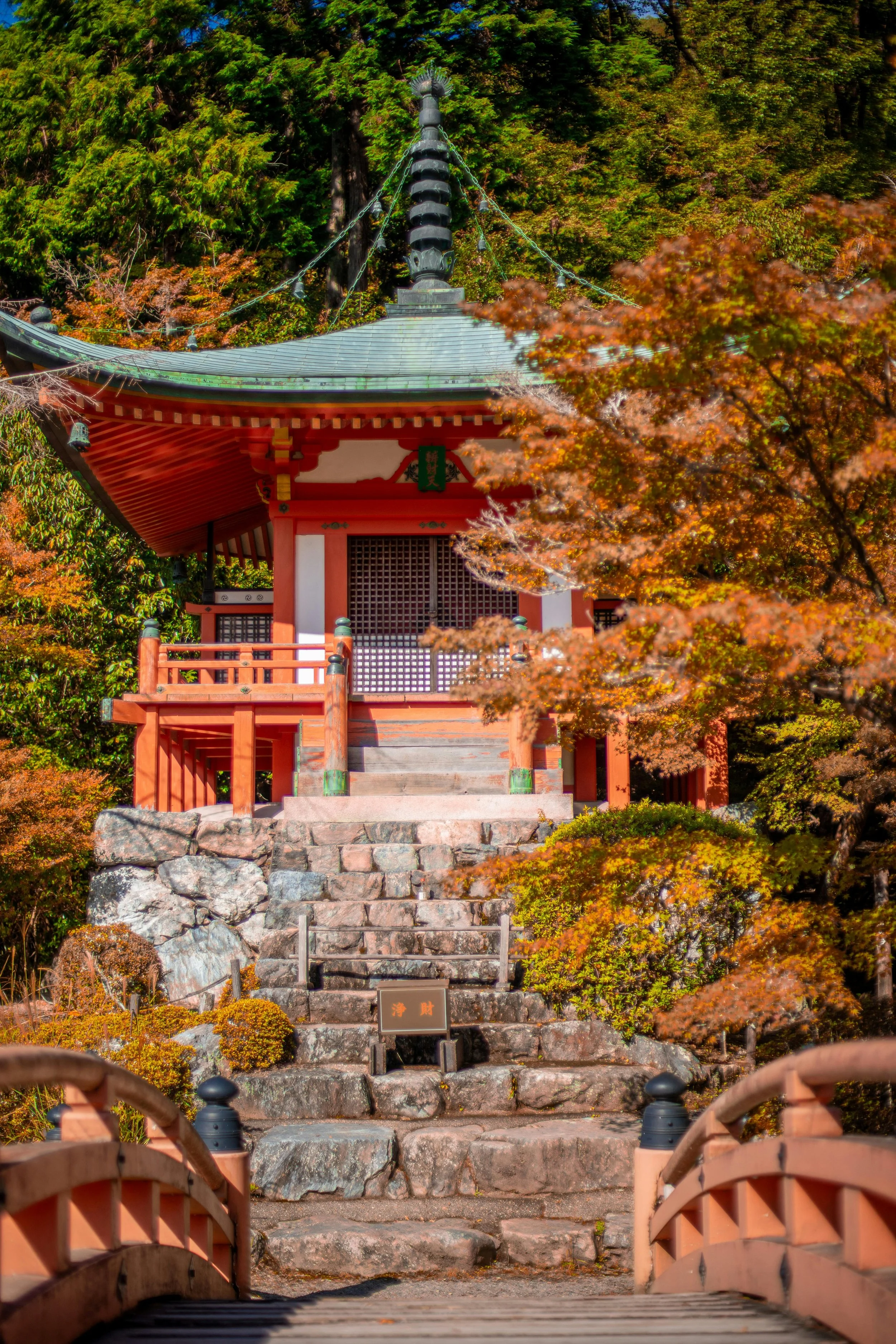 A traditional Japanese temple or shrine elevated on stone steps, with a small bridge in the foreground and autumn-colored trees surrounding it.