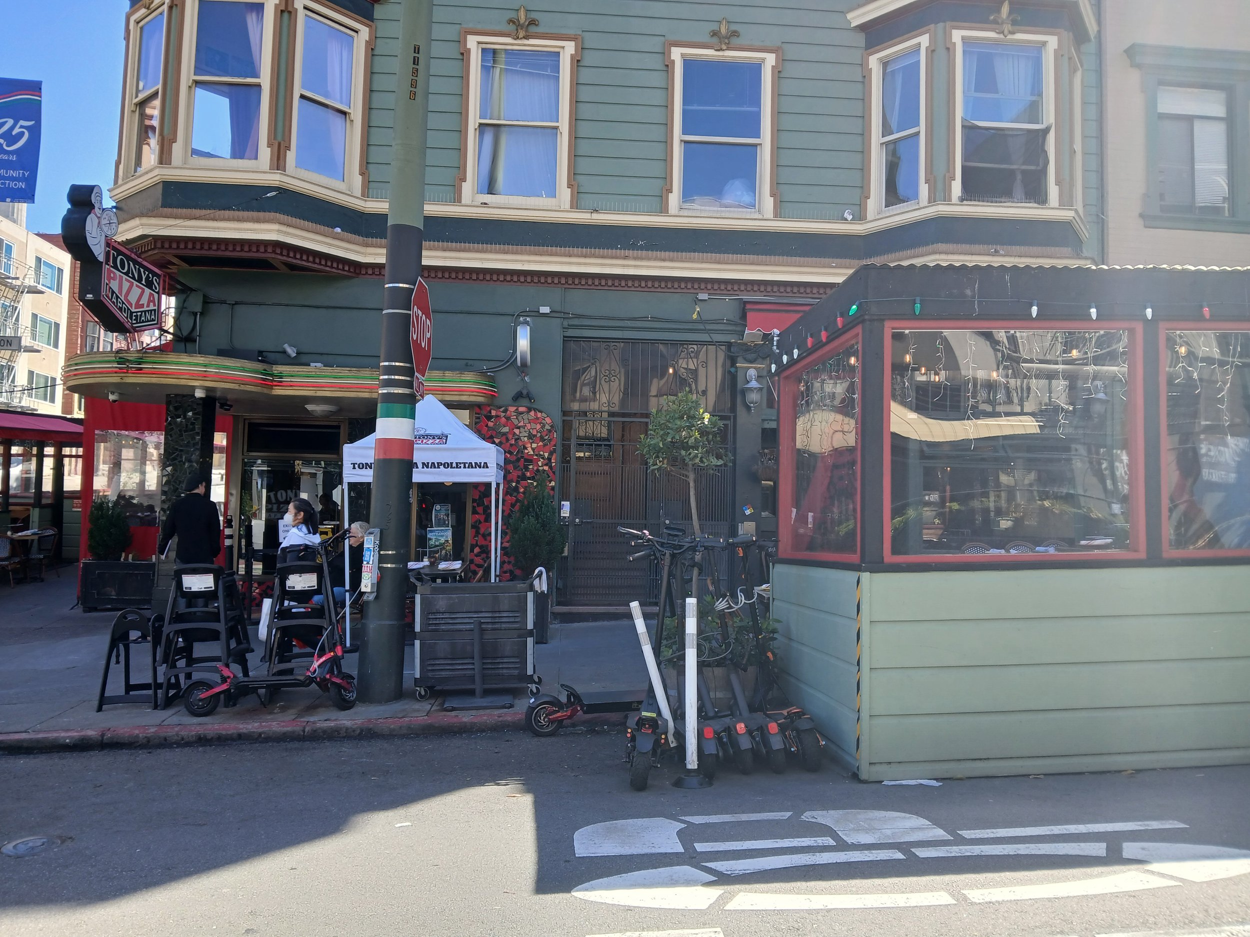 Street view with a green building housing Tony's Pizza Napolitana, outdoor seating, bicycles, and a small market stand with tools, and scooters parked outside.