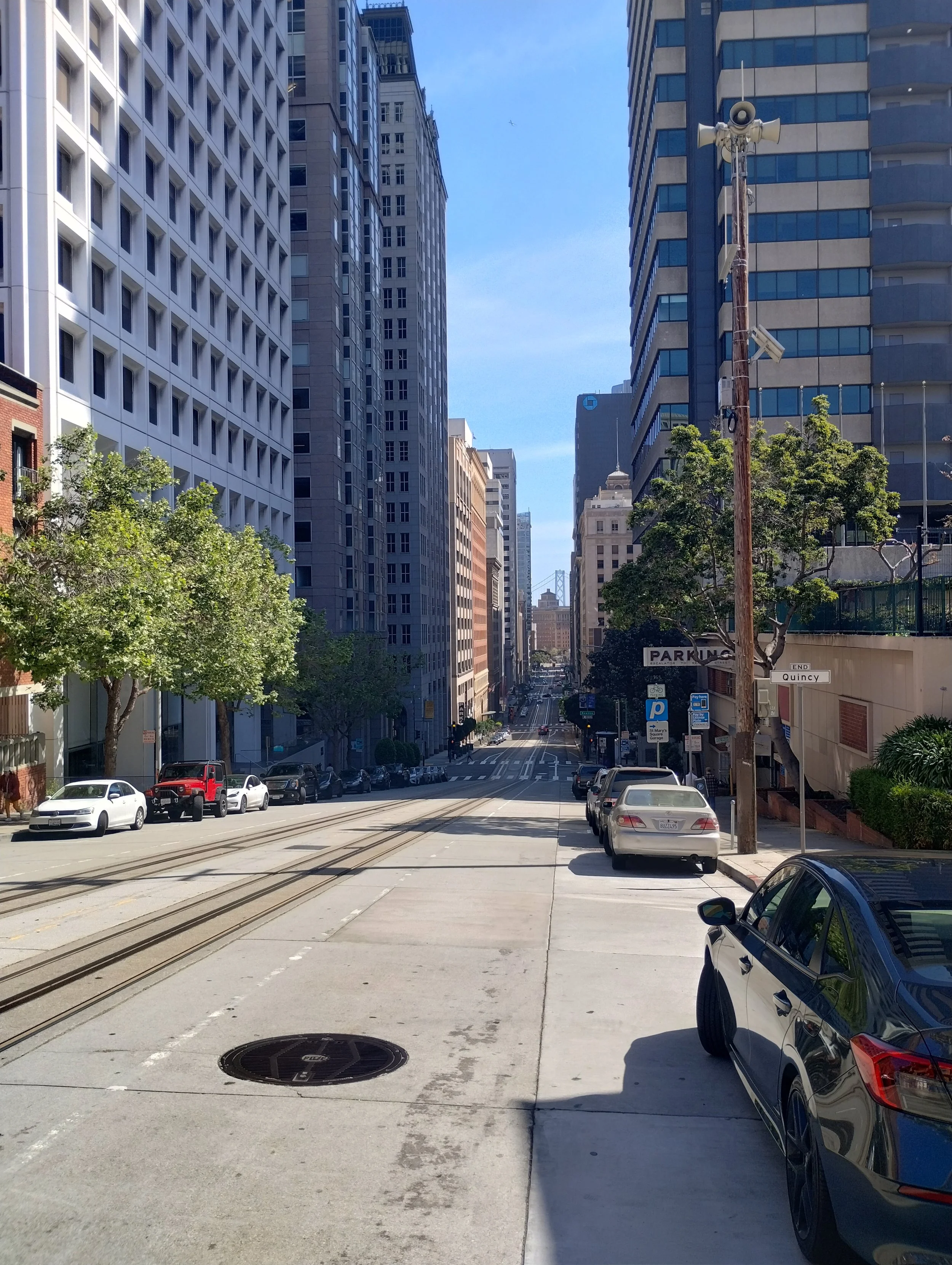 City street view with parking lot, parked cars, tall buildings, trees, a street sign indicating 'Quincy' and 'Park Ave', and a clear blue sky.