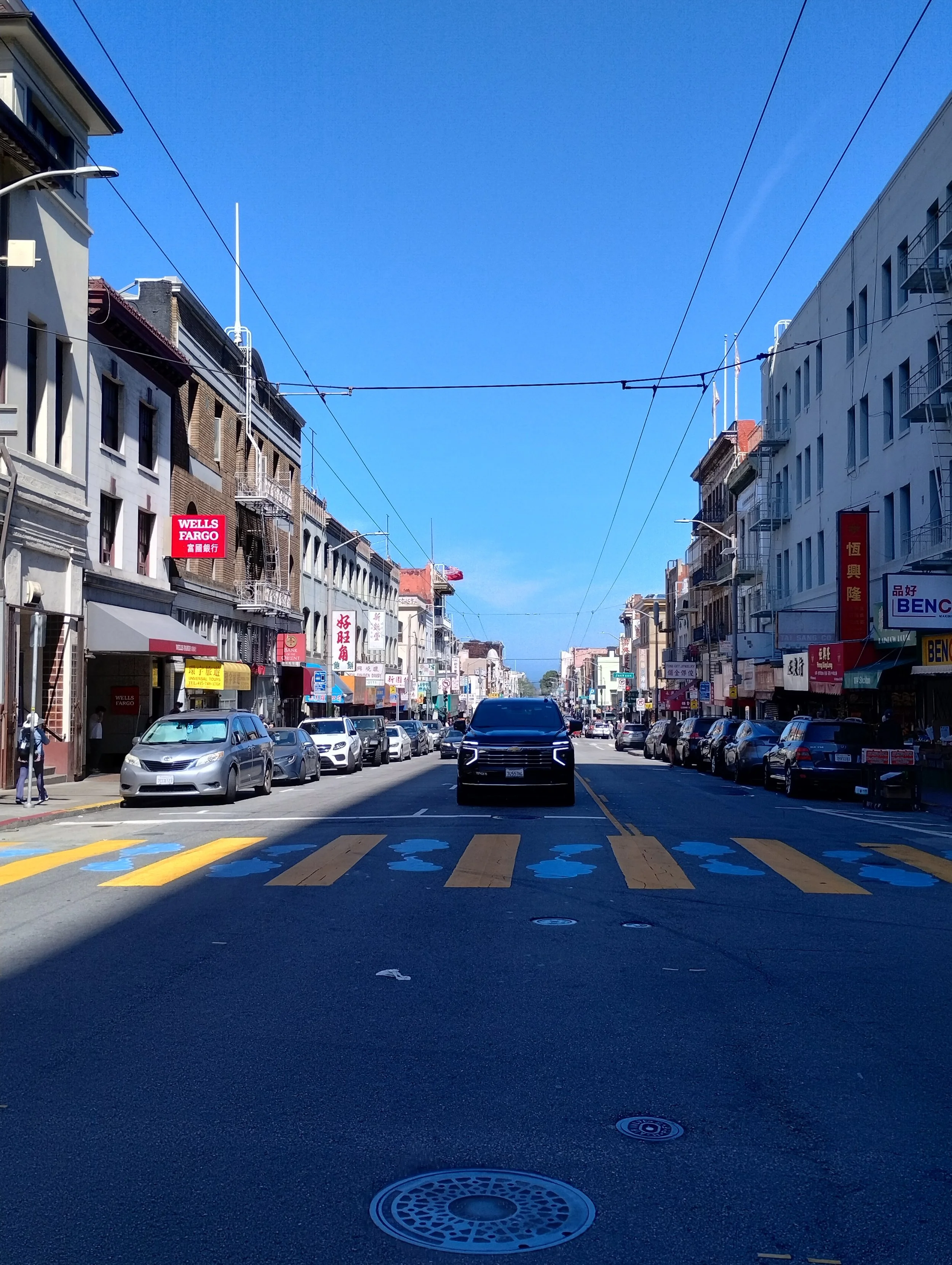 A city street with cars parked along both sides, buildings with signs in multiple languages, and a clear blue sky overhead.