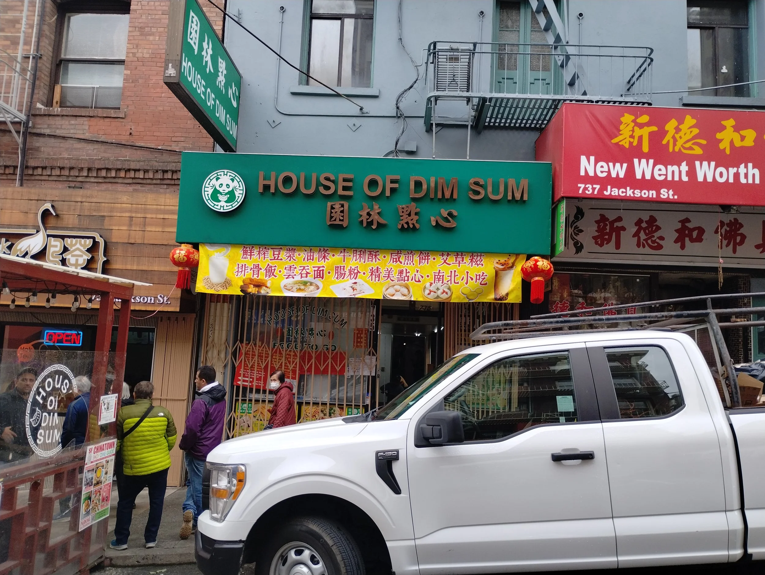 The image shows a busy street scene in front of a restaurant called 'House of Dim Sum' with a green sign and logo. Several people are visible, some waiting outside, and a white truck is parked in front of the restaurant. There are other signs in Chinese and English, including a red sign for 'New Went Worth' at 737 Jackson St.