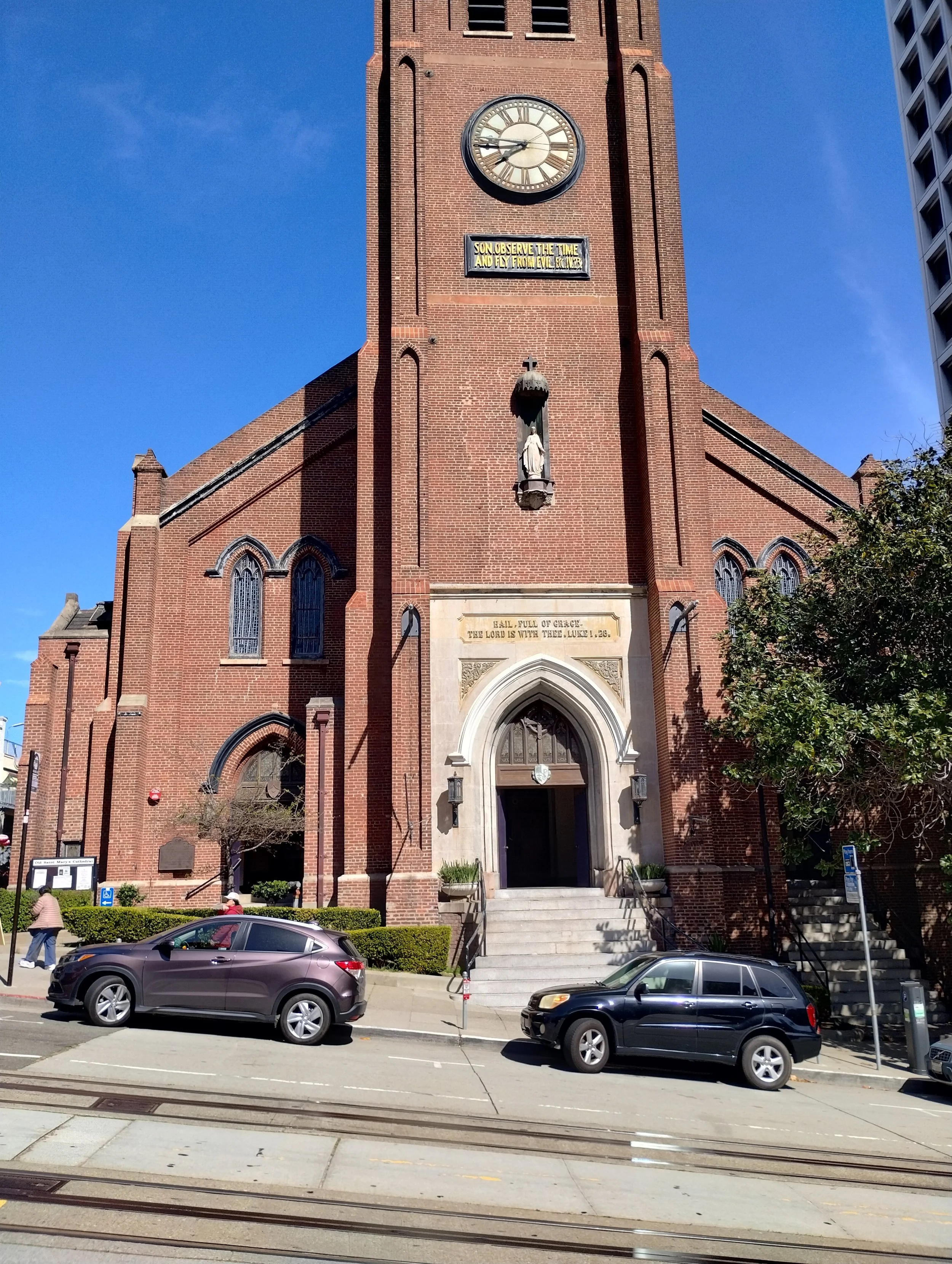 The front view of a red brick church with a clock on its tower showing the time as 9:50. There is a statue of Jesus Christ in a niche above the entrance, which is marked with a cross on top. Several parked cars are visible in front, with pedestrians walking on the sidewalk. The sky is clear and blue.