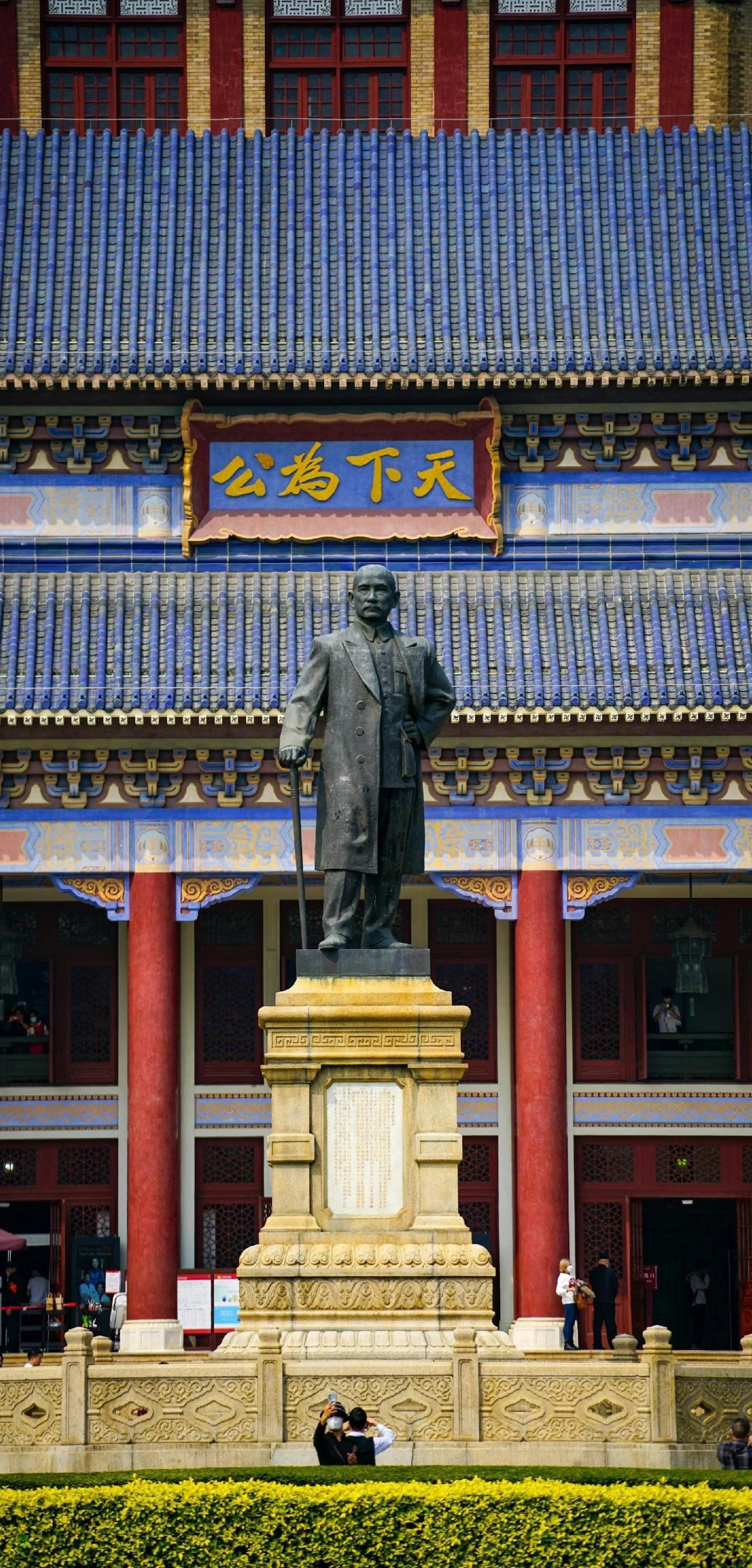 A statue of a man standing outside a traditional Chinese building with a blue tiled roof and red columns. The man in the statue holds a cane and is dressed in a long coat. The building has a sign with Chinese characters in yellow. There are people near the entrance, and the scene includes a well-maintained yellow flower bed in the foreground.