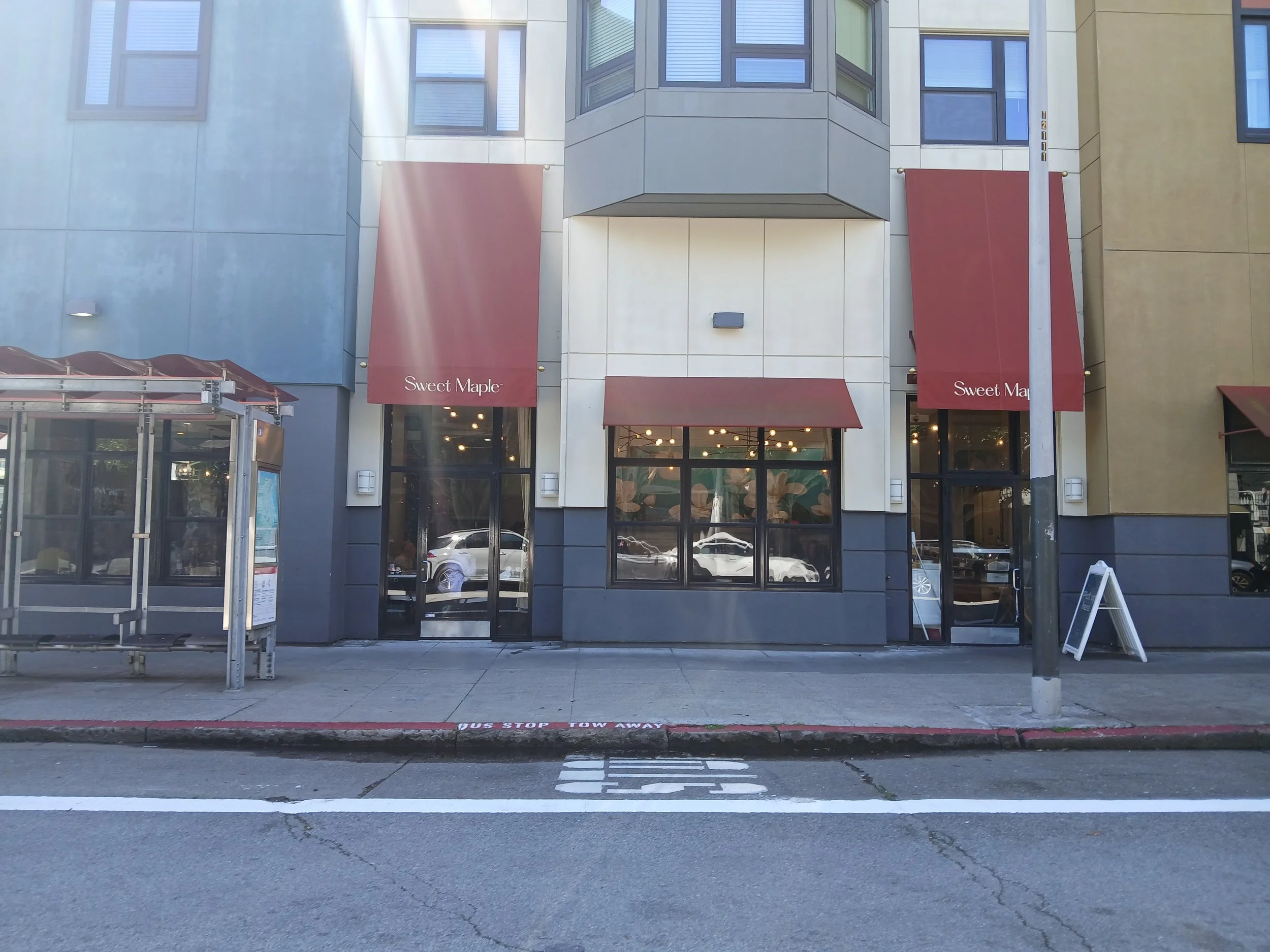 Front view of a commercial building with large glass windows, red awnings labeled 'Sweet Maple', and a sidewalk with a parking lane.