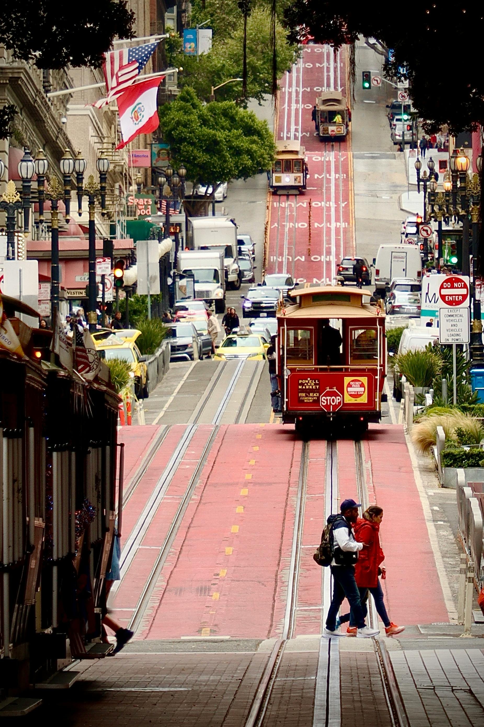 A street scene with a historic trolley car traveling on red tram tracks through a city. Pedestrians are walking along the sidewalks, and decorative street lamps line the street. There are flags, including the American and Peruvian flags, hanging from buildings. Vehicles are parked along the sides, and a traffic light is green. The street has buildings with storefronts and trees, suggesting a lively urban area.
