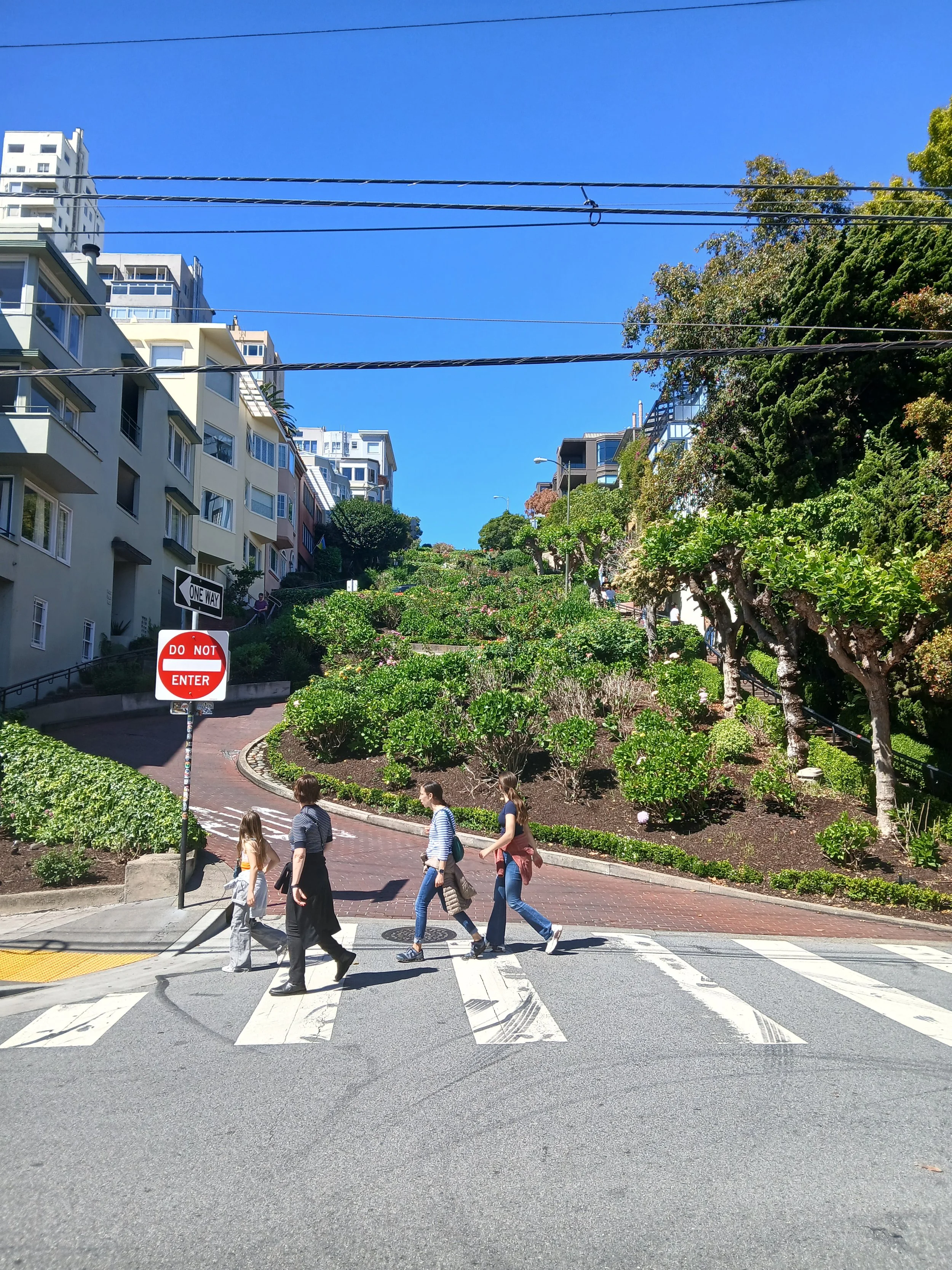 People crossing a street at a crosswalk in an urban area with residential buildings and greenery in the background on a sunny day.