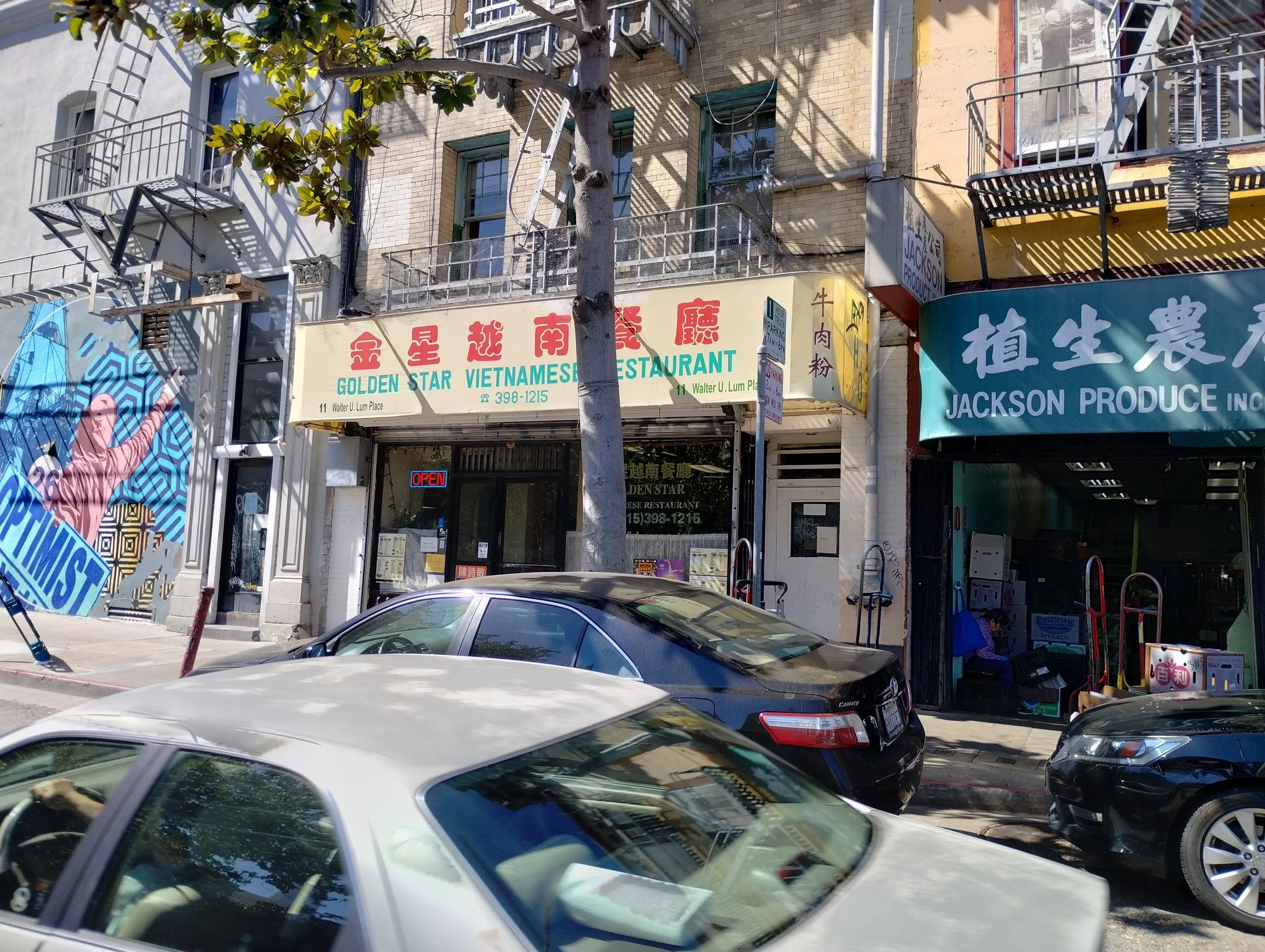 Street scene with storefronts, parked cars, a tree, and building fire escapes. Visible signs for a Vietnamese restaurant and produce store.