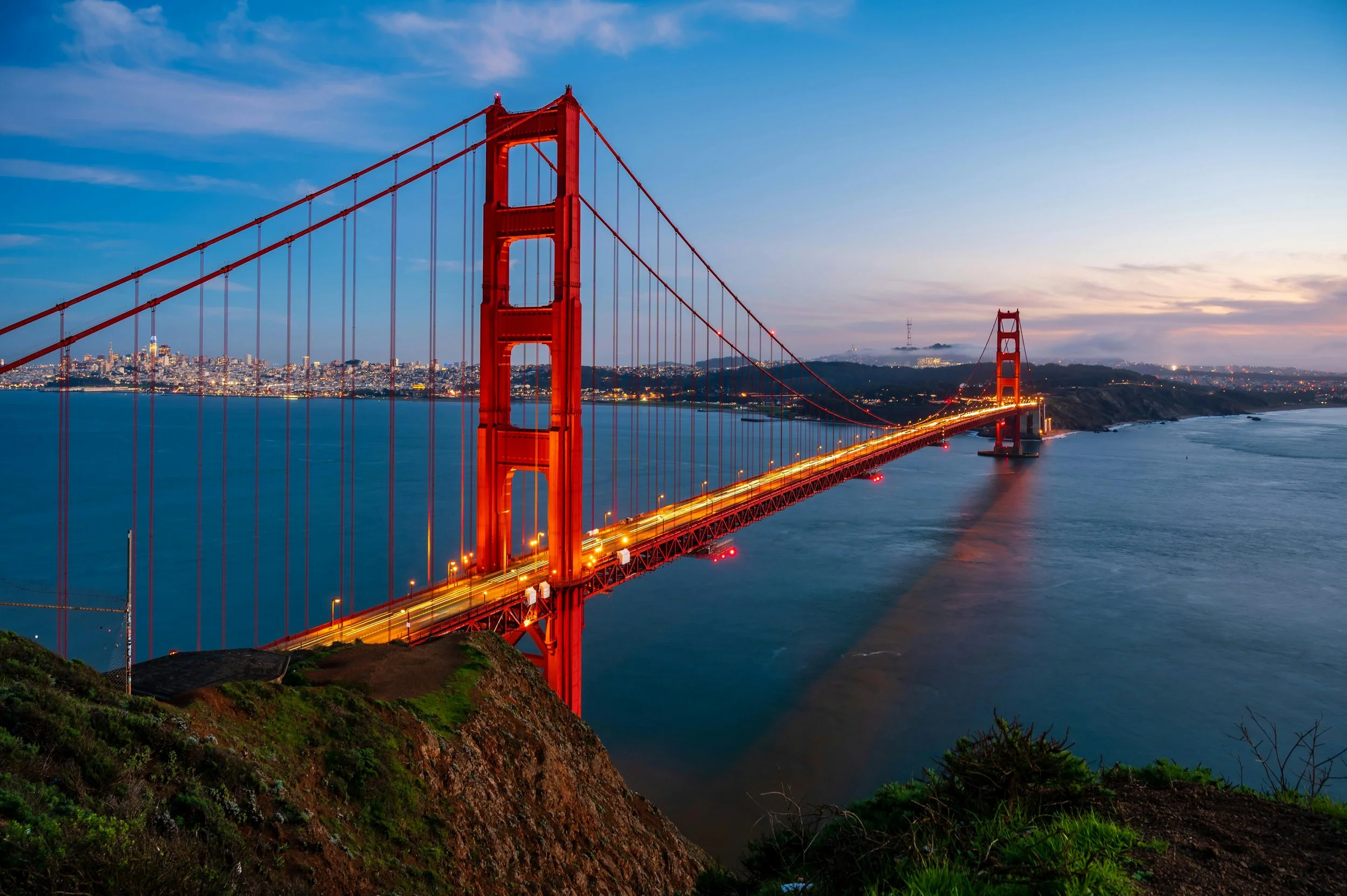 The Golden Gate Bridge in San Francisco at dusk, illuminated with lights, with the city skyline in the background and calm waters below.