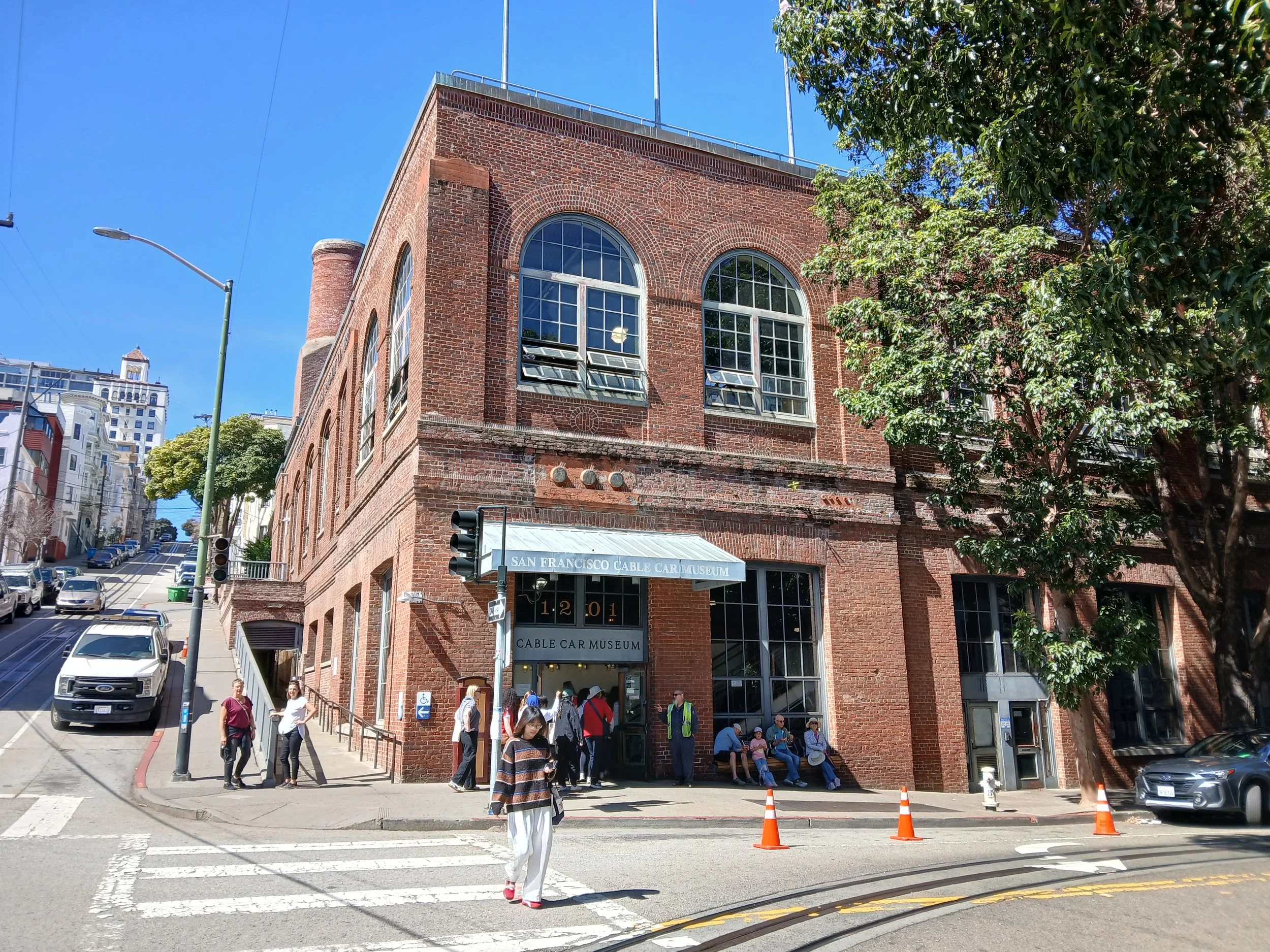 The image shows a brick building with large arched windows. There are several people sitting and standing outside the entrance, which has a sign that reads 'SAN FRANCISCO CABLE CAR MUSEUM'. Some people are walking across the street, and there are parked cars along the lane. The sky is blue, and a traffic light and streetlamp are visible. Trees partially obscure the right side of the building.