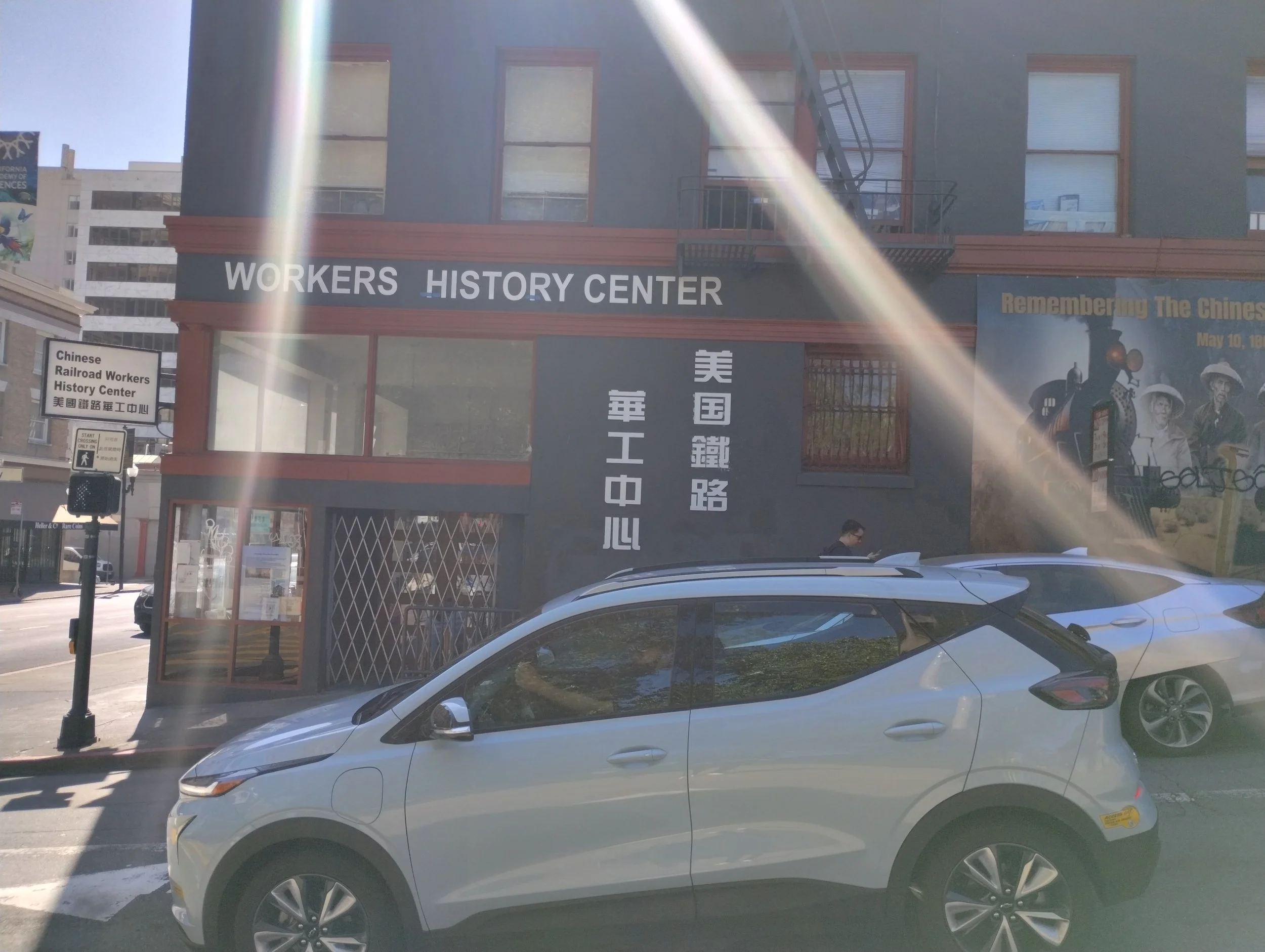 Street view showing a building with a sign that reads 'Workers History Center,' with cars parked in front and a person walking nearby.