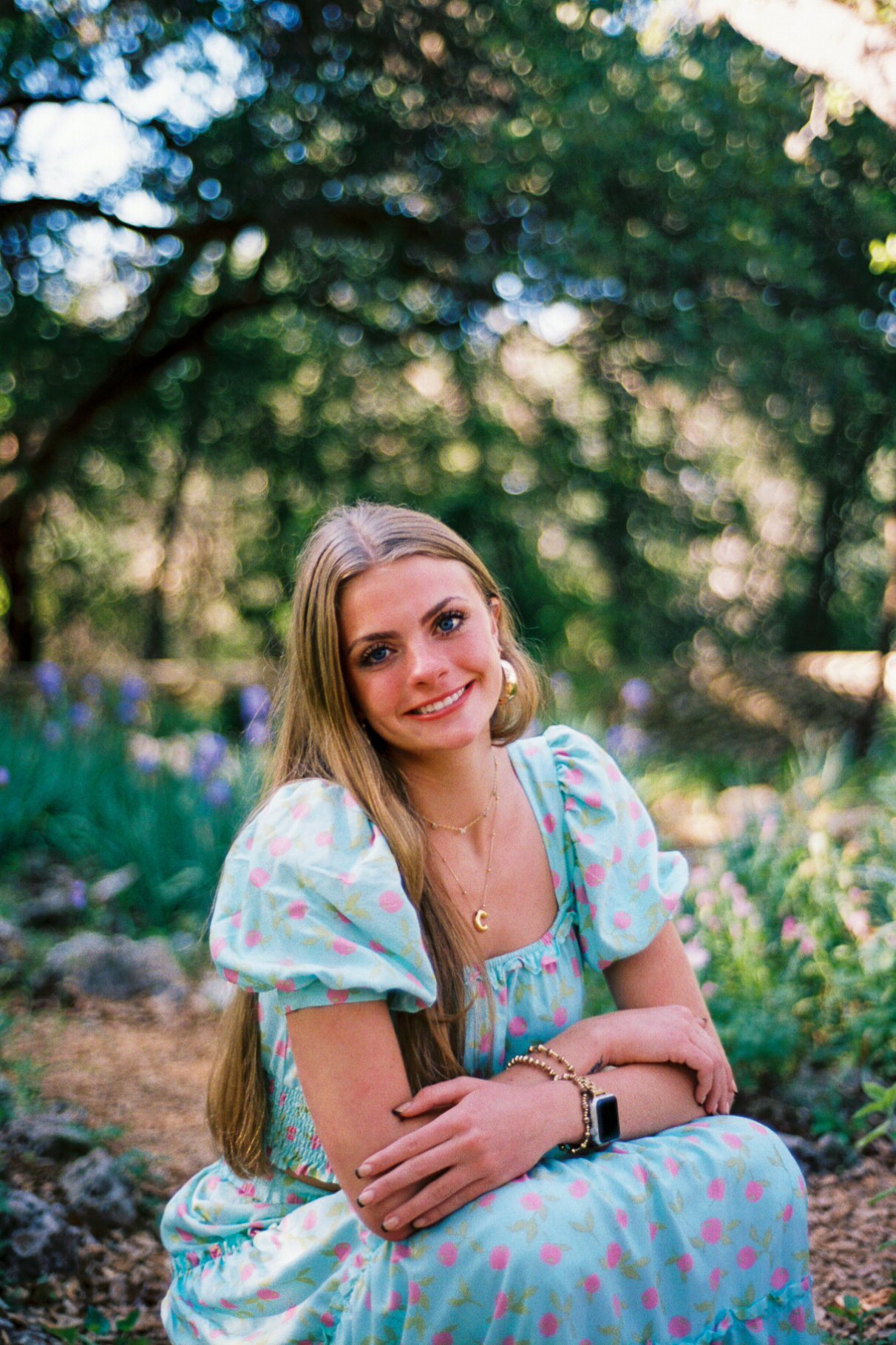 A young woman with long blonde hair and blue eyes, smiling, sitting outdoors on the ground with trees and greenery in the background. She is wearing a light blue floral dress with puffed sleeves, gold jewelry, and a smartwatch.
