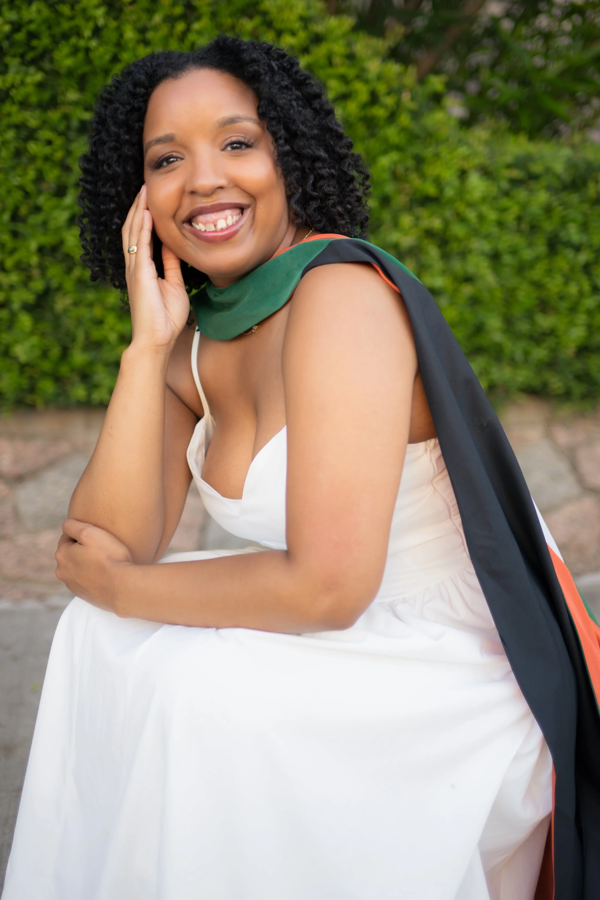 A woman smiling outdoors, wearing a white dress and a sash, with a black backpack over her shoulder, posing in front of green bushes.