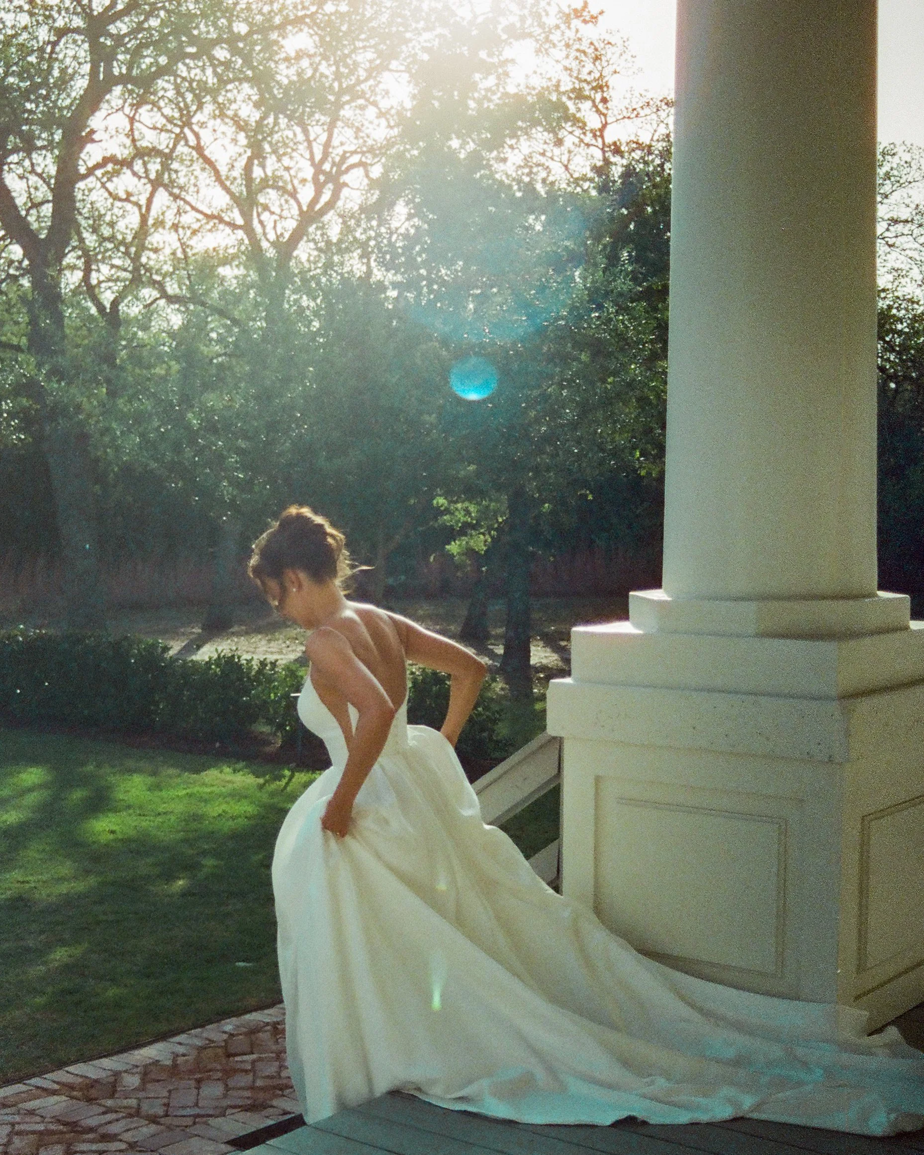 A woman in a white gown playing a white grand piano outdoors on a brick and wood deck, with trees and sunlight in the background.
