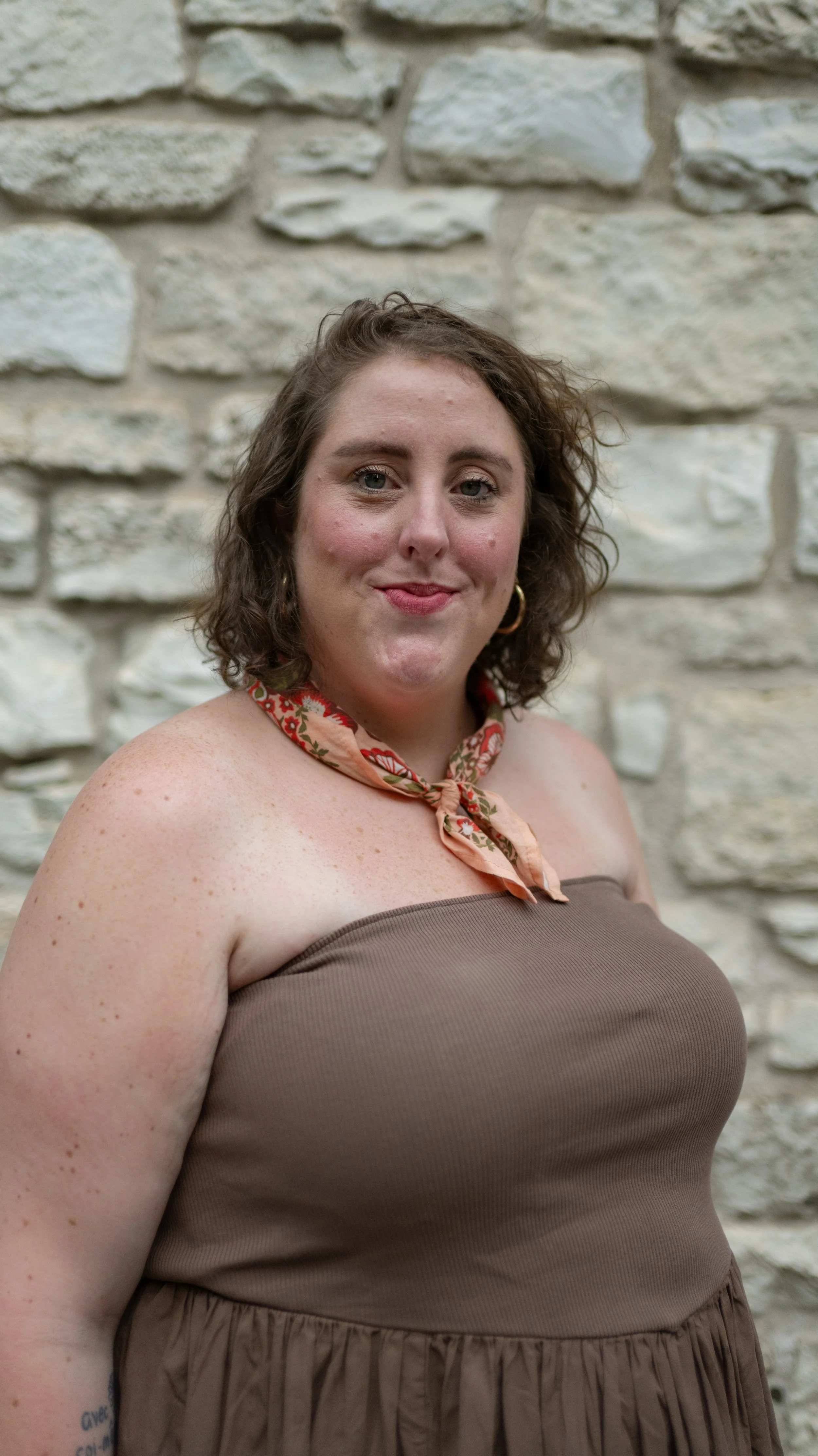 A woman with curly brown hair wearing a brown strapless dress and a pink floral scarf tied around her neck, standing in front of a stone wall.