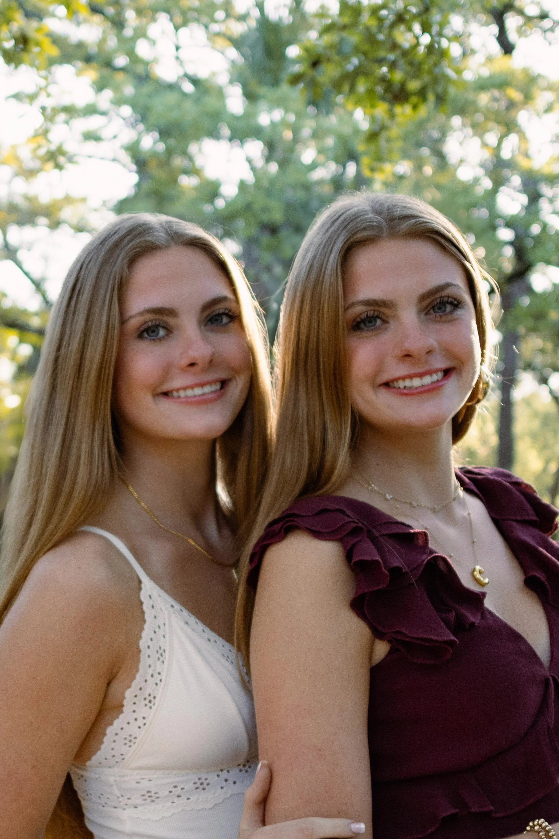 Two young women with long, light brown hair and blue eyes standing outdoors in a natural setting with trees and sunlight in the background. They are smiling and dressed in sleeveless tops, one in white and the other in maroon, wearing jewelry.