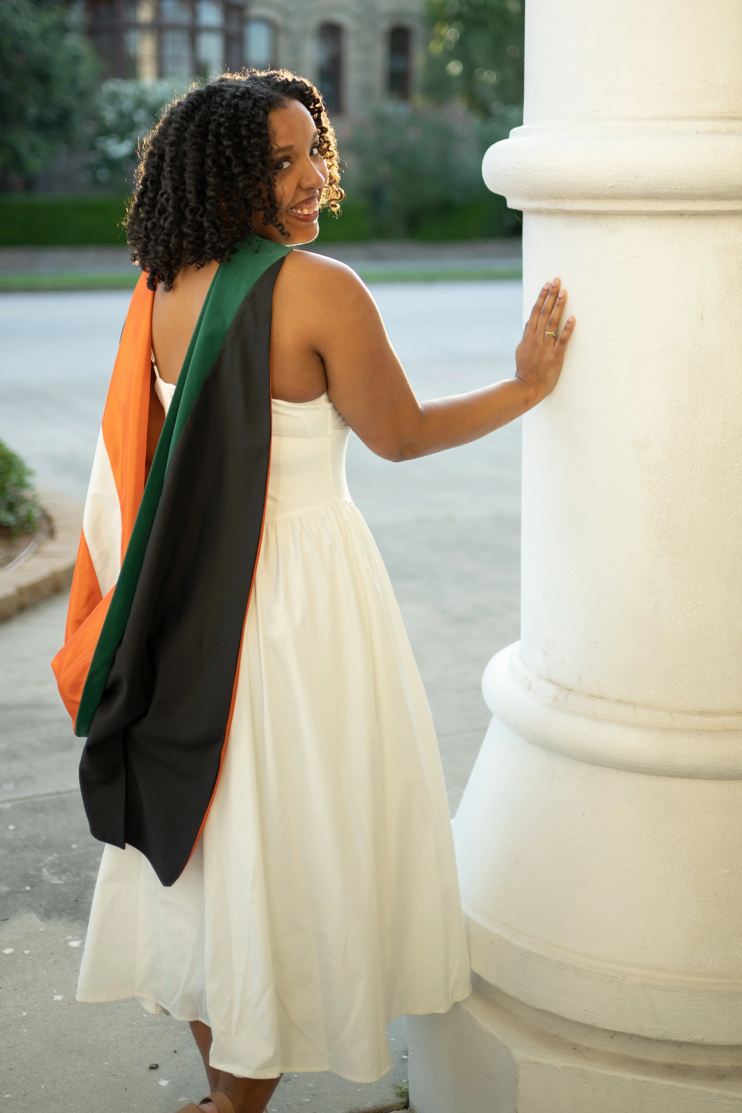 A woman with curly hair and medium skin tone in a white dress touching a white column of a building, with a black, orange, green and white flag draped over her shoulder, outdoors during daytime.