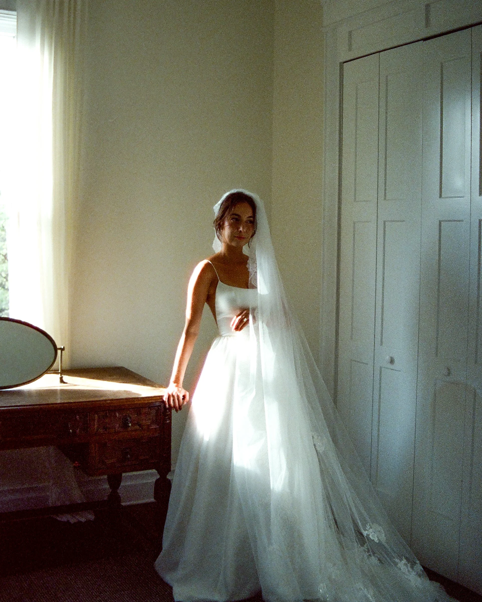 A bride in a white wedding dress with a veil standing beside a wooden dresser in a room with white walls and a door, sunlight coming through a window.