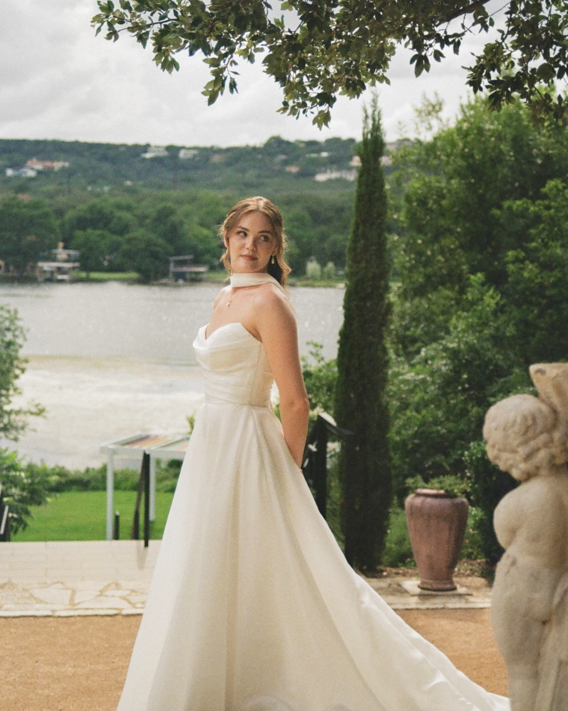 A woman in a white dress appears to be at an outdoor event near a body of water, with trees and greenery in the background.