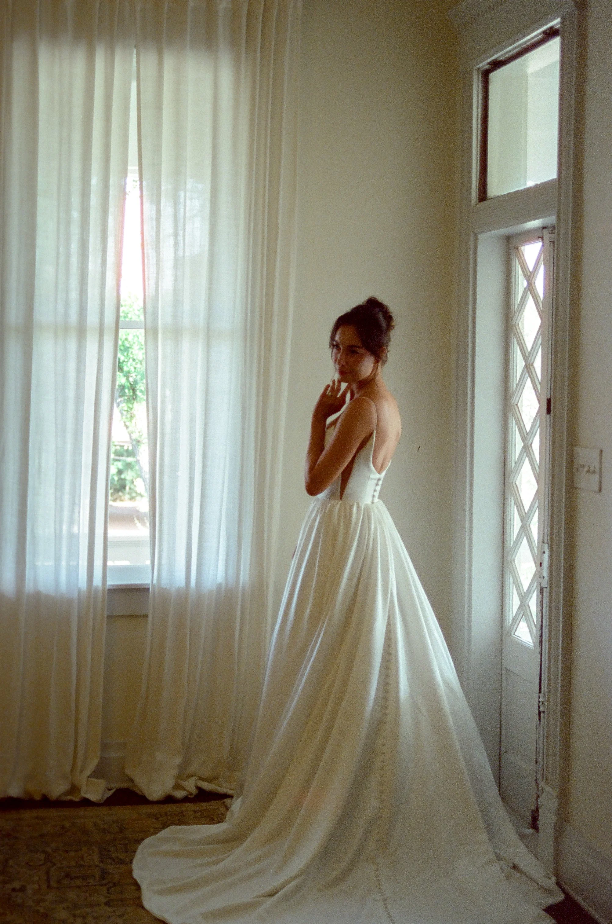 A woman in a cream dress stands indoors near large windows with white curtains, natural light illuminating a vintage wooden door with diamond-shaped panes, and a richly patterned carpet on the floor.