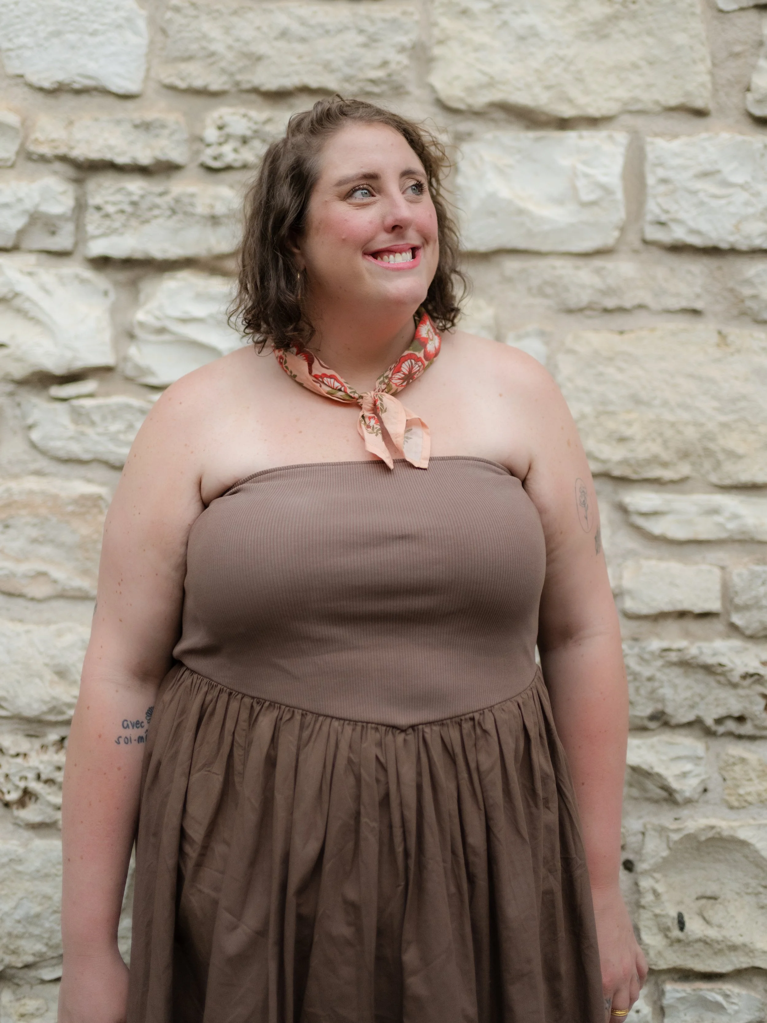 A woman with curly brown hair smiling, wearing a brown dress with a floral scarf around her neck, standing in front of a stone wall.