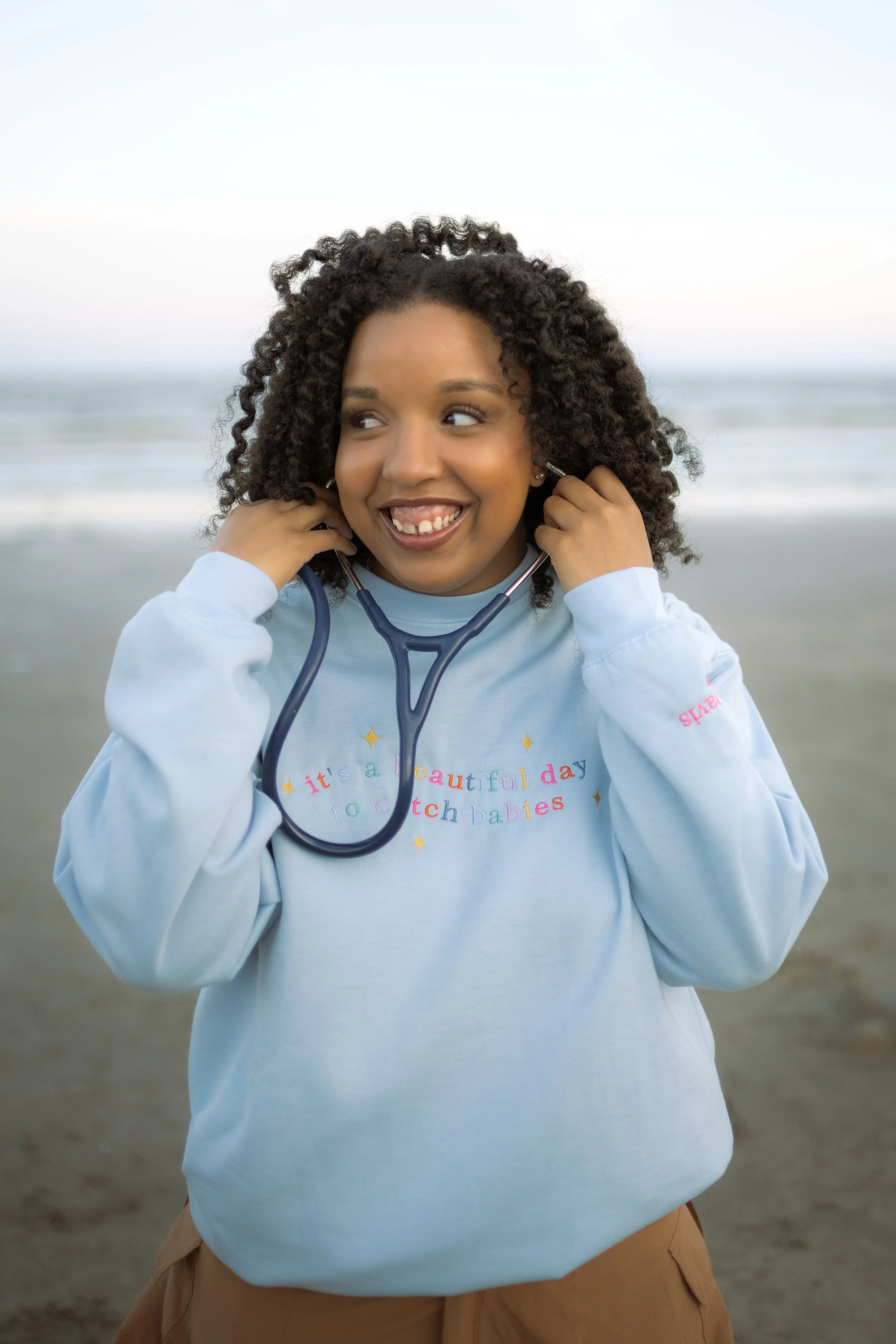 A smiling young girl with curly hair at the beach, holding a stethoscope around her neck.