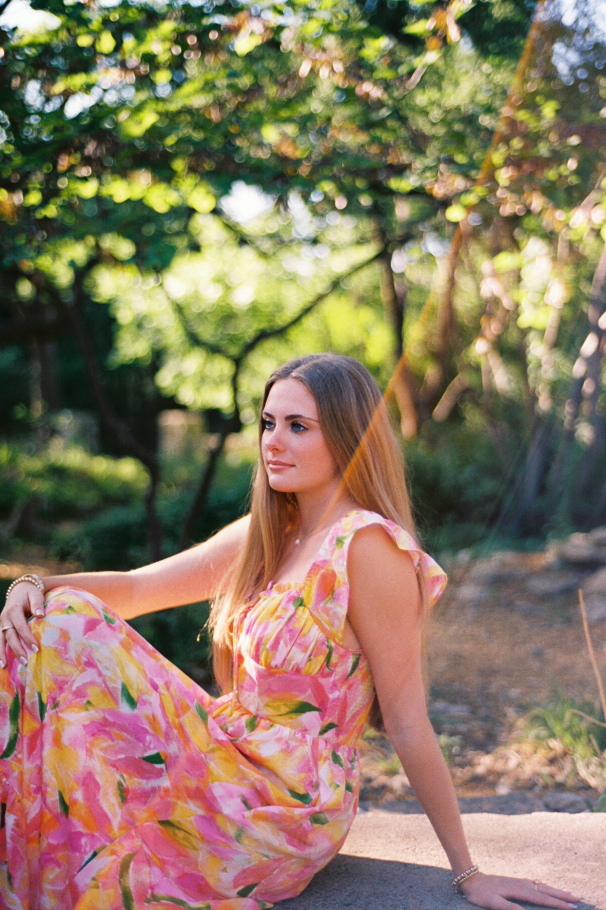 Young woman in a colorful yellow-pink floral dress sitting on the ground outdoors in a lush green park.