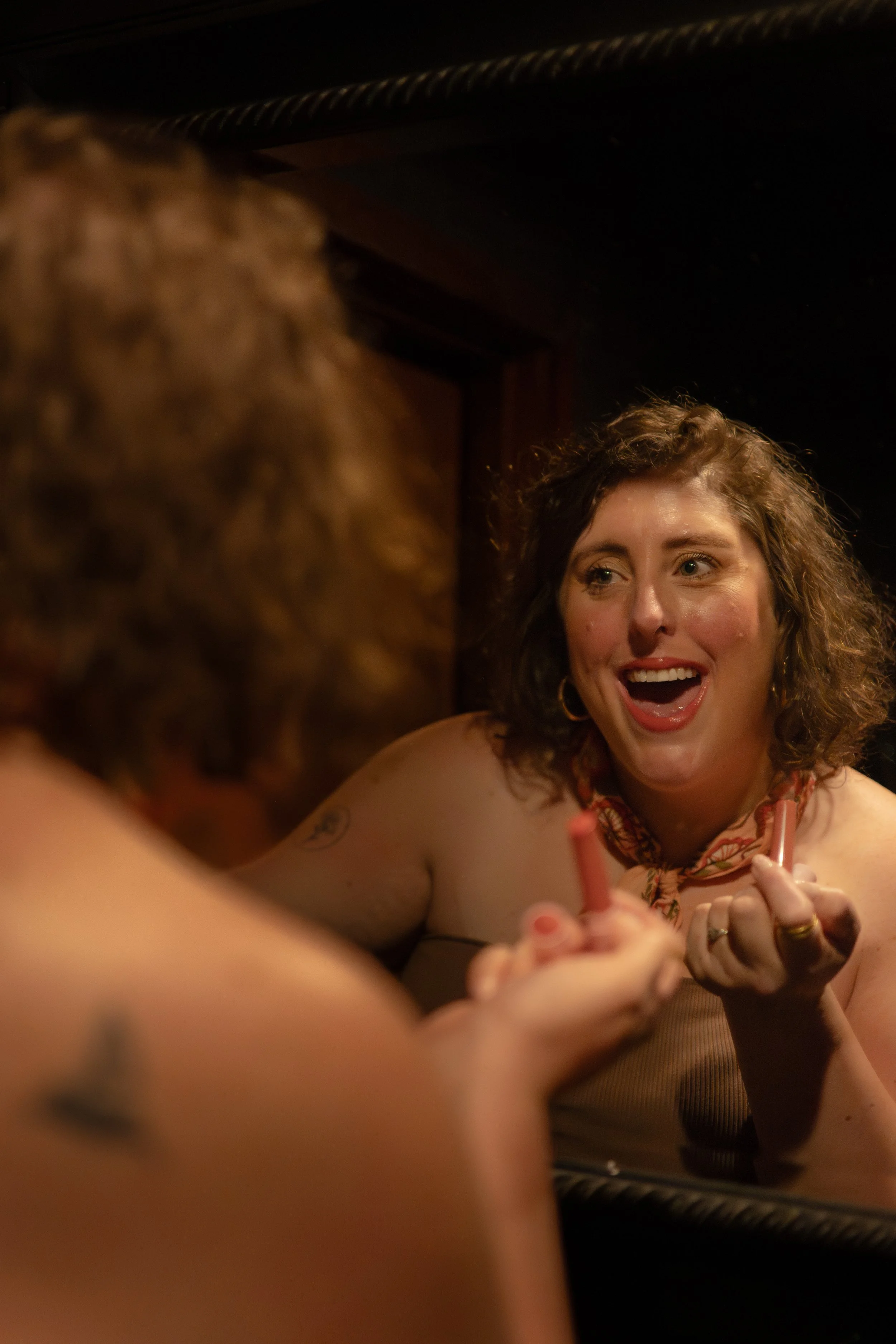 A woman with curly hair and a big smile looking at her reflection in a mirror, holding a makeup brush.