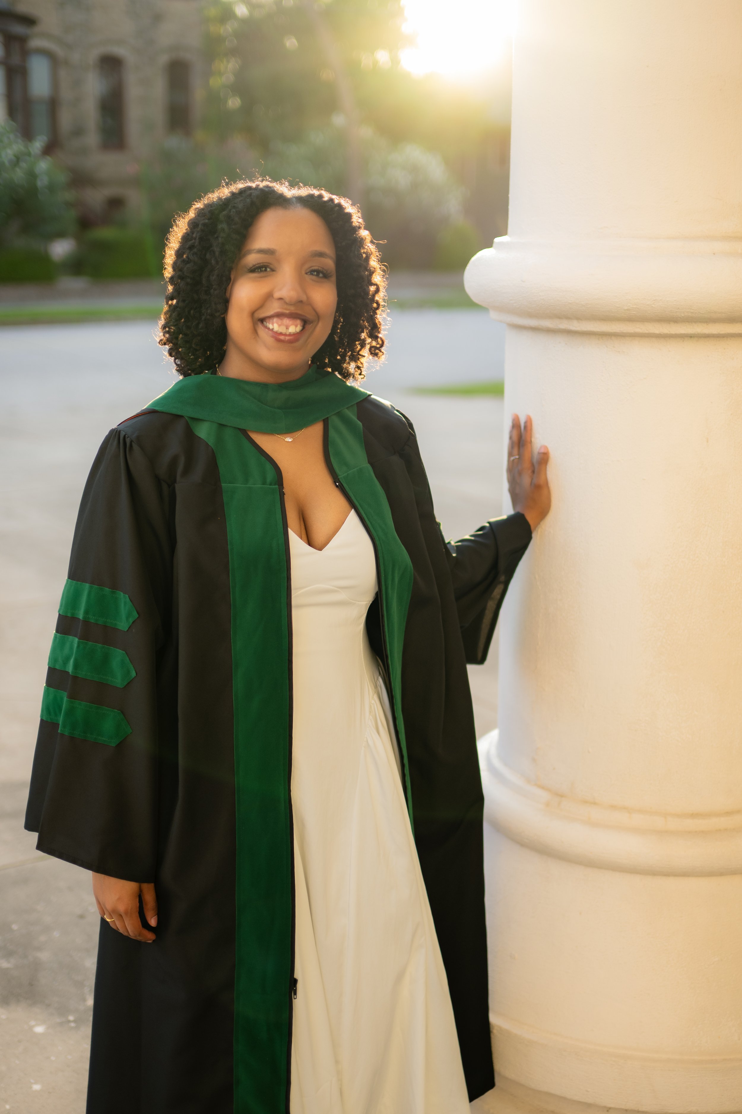 A smiling woman dressed in a graduation gown and cap, standing outdoors in front of a large column, with a building and trees blurred in the background during sunset.