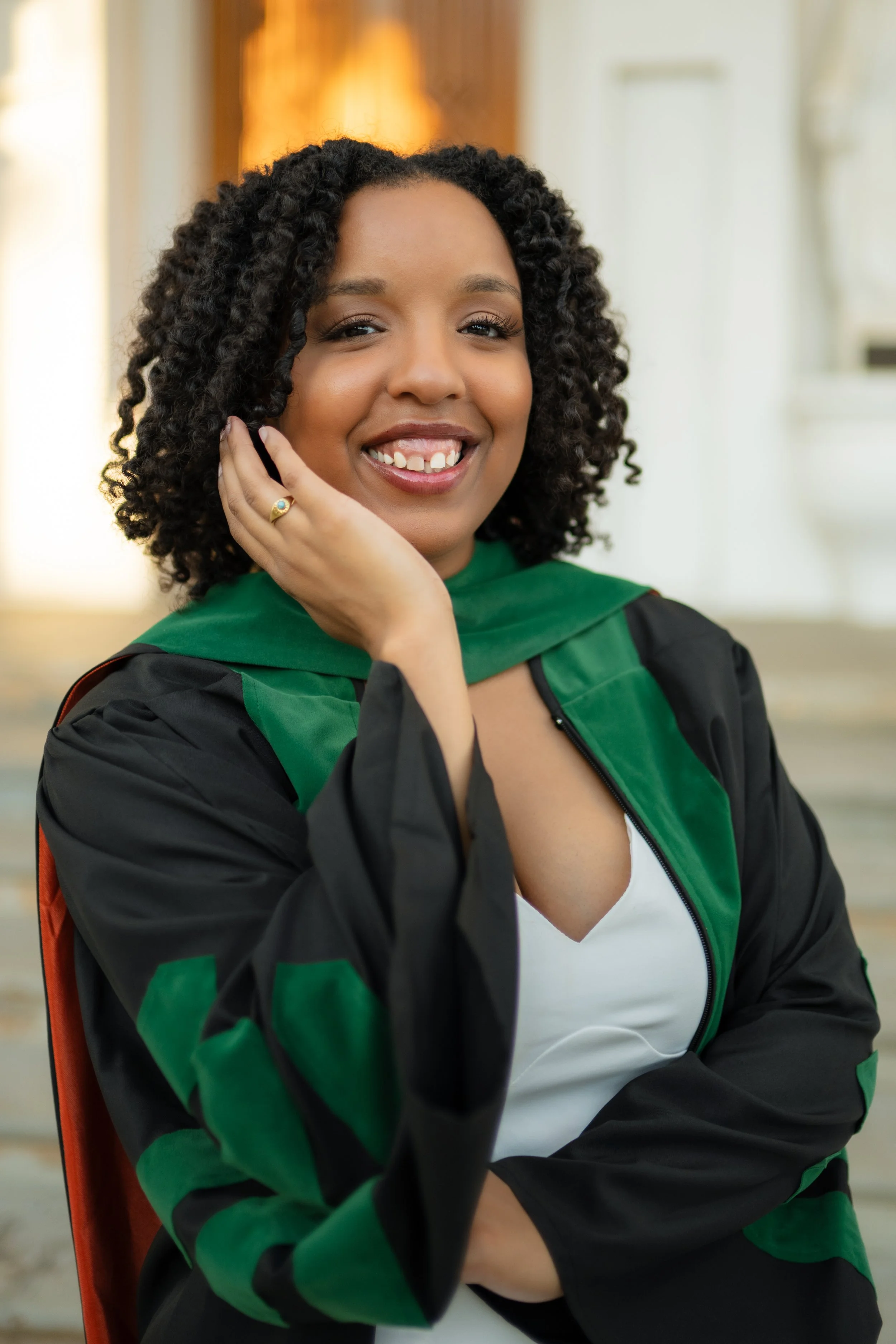 A young woman with curly black hair and missing front teeth smiling, wearing a black and green graduation gown and white shirt, sitting on an orange chair in an indoor space.