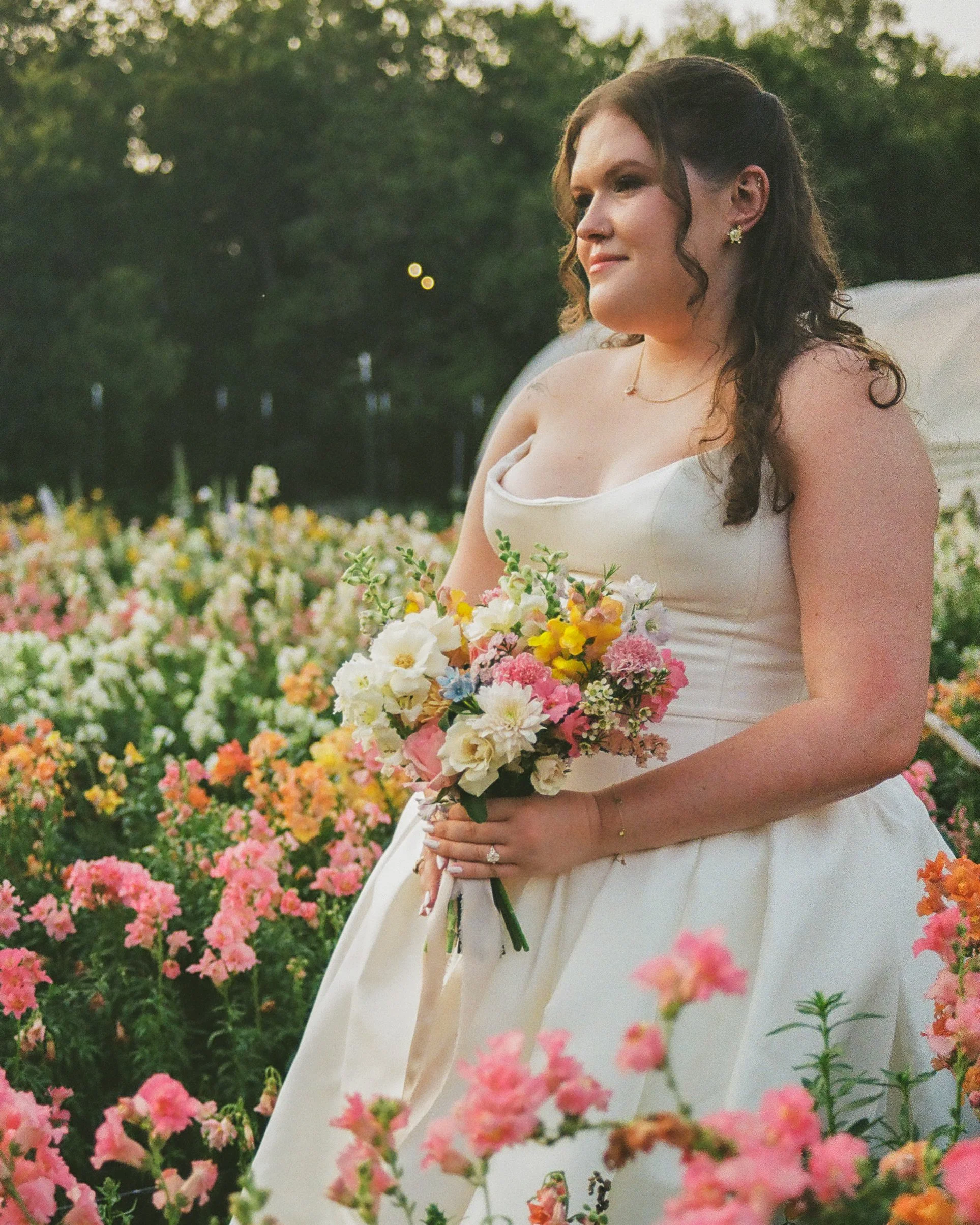 A bride in a white dress holding a bouquet of colorful flowers, standing outdoors among pink, white, and yellow blossoms with greenery in the background.
