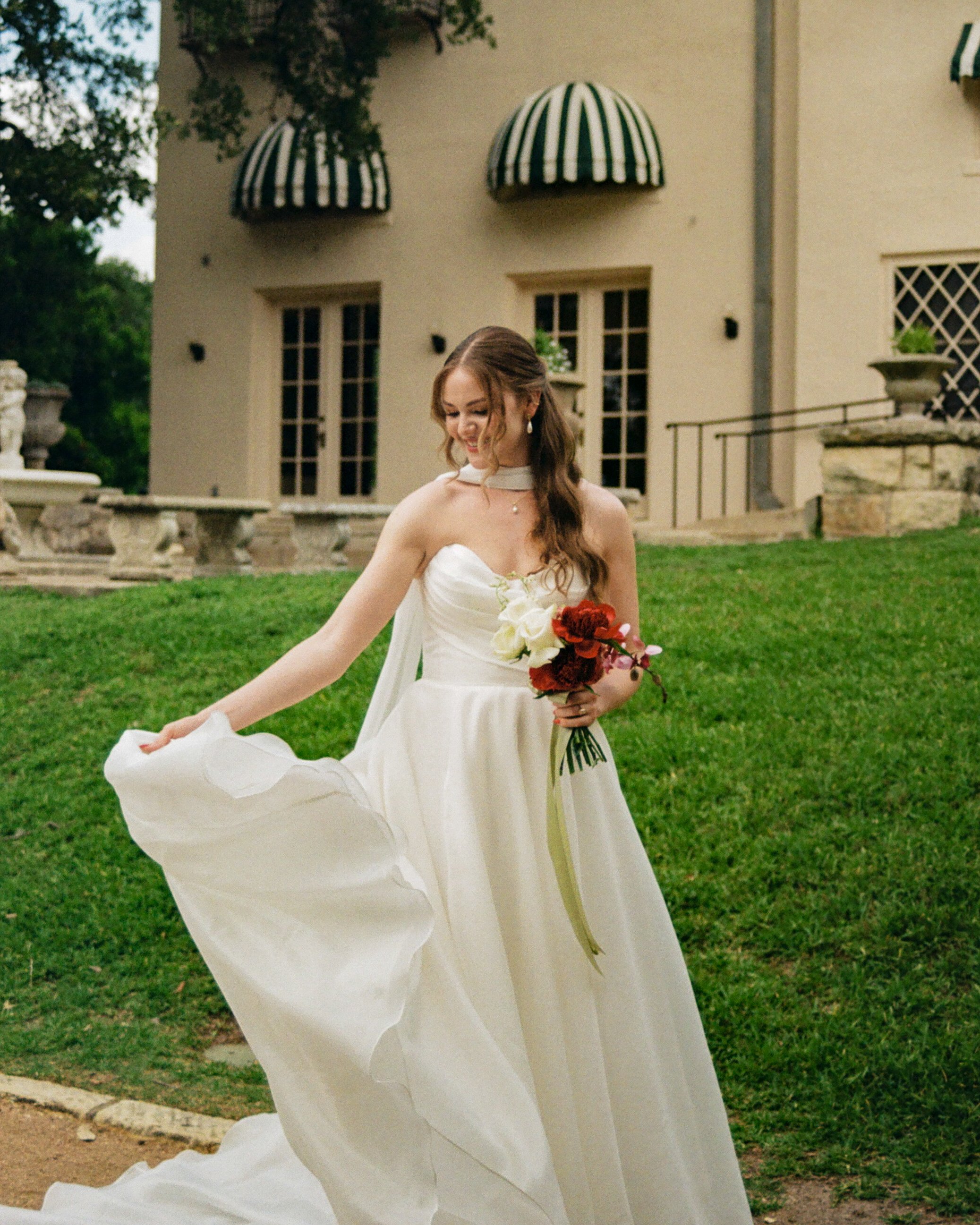 A woman in a white dress holding a bouquet of flowers stands outdoors on a grassy area in front of a beige building with a black and white striped awning.