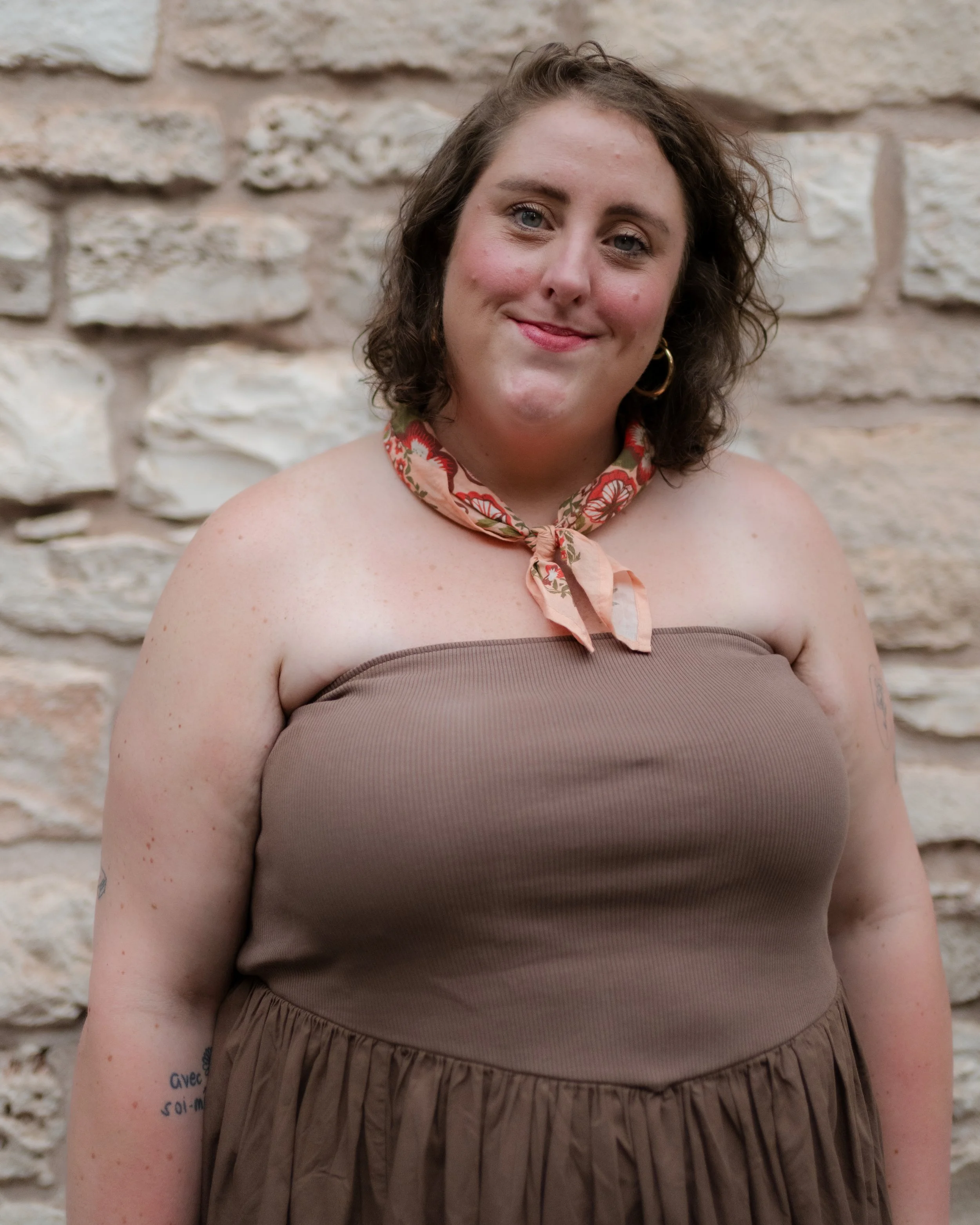 A woman with curly brown hair wearing a brown strapless dress, a floral scarf around her neck, and hoop earrings, standing in front of a stone wall.