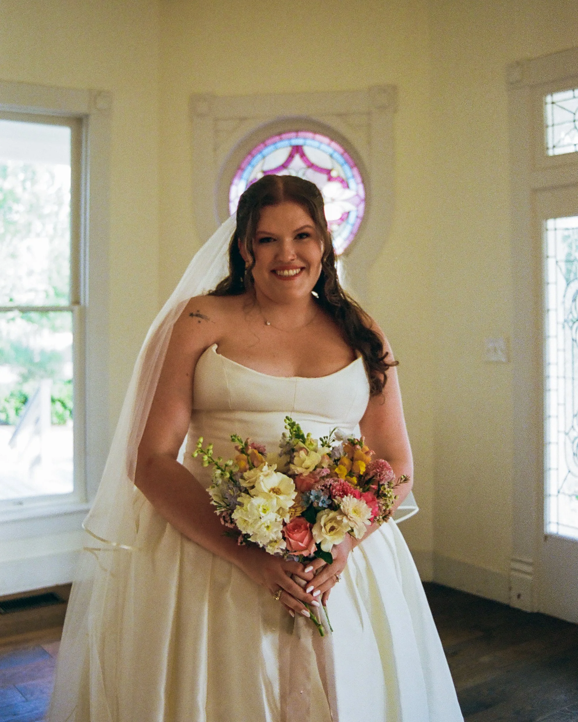 A smiling bride with long brown hair in a wedding dress holding a colorful bouquet of flowers inside a church with stained glass windows.