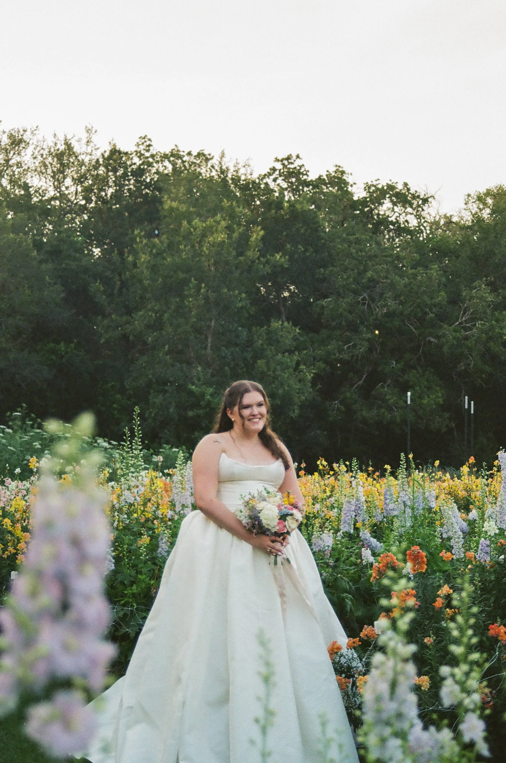 A woman in a white wedding dress holding a bouquet of flowers, standing in a colorful flower garden with trees in the background.