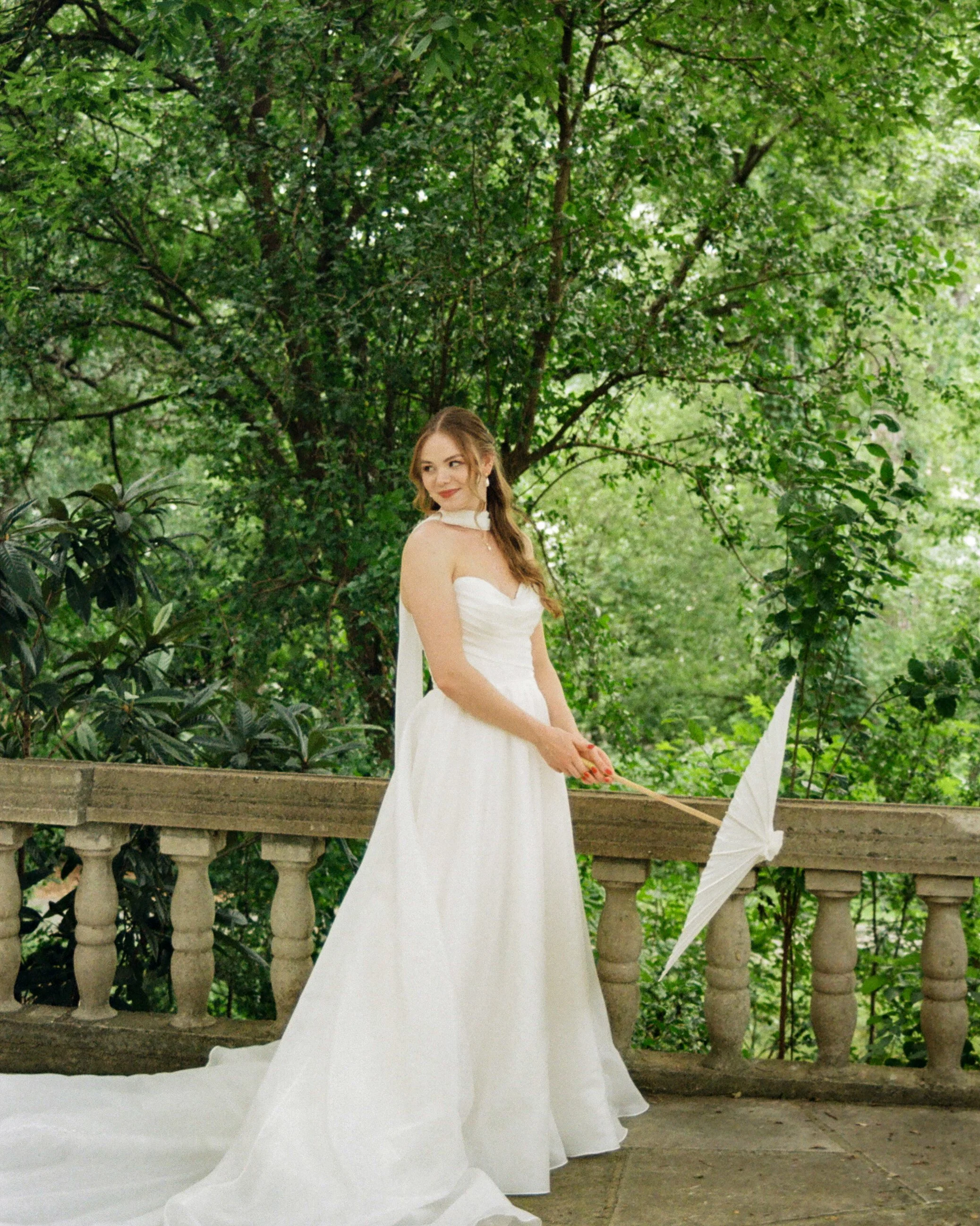 A woman wearing a white dress with a sheer cape, standing outdoors near a decorative stone balustrade and green foliage.