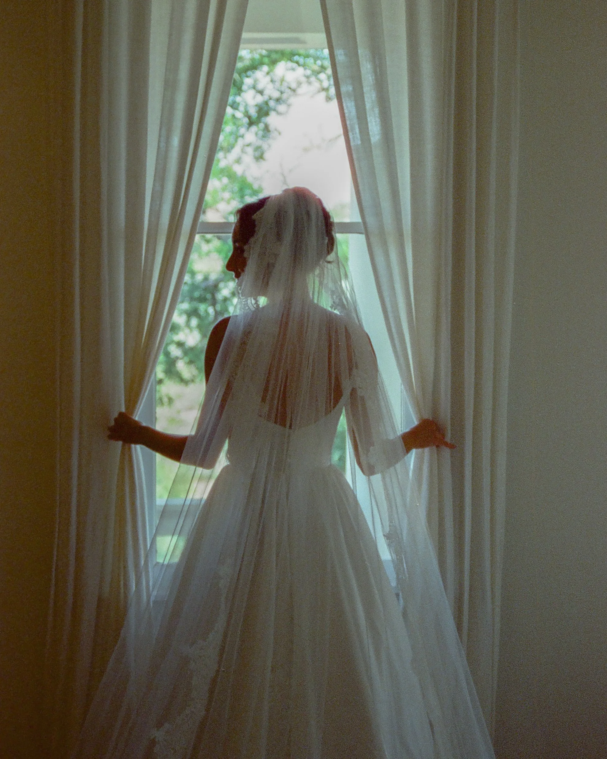 Woman in wedding dress standing behind sheer curtains, looking out the window to green trees outside.