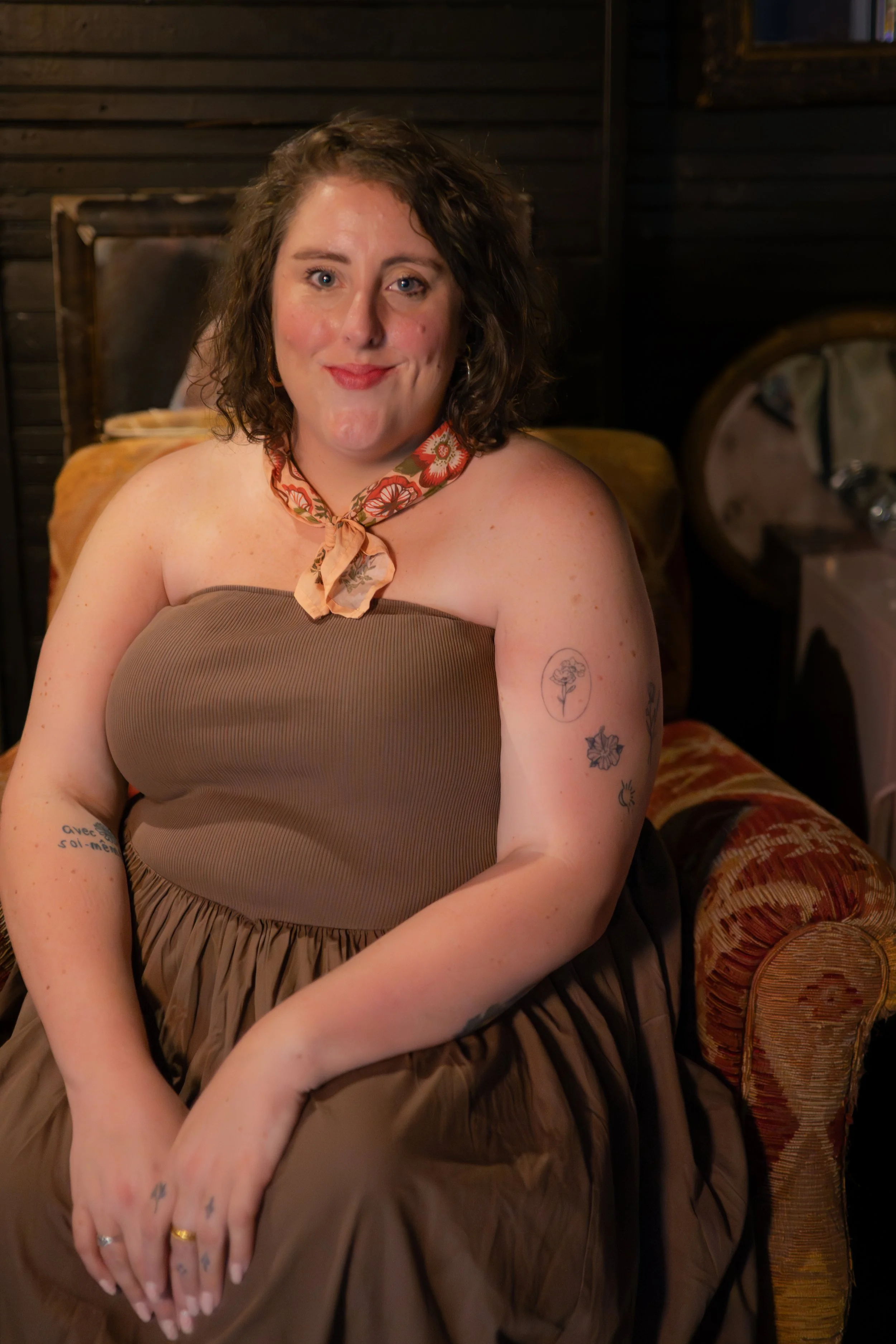 A woman with curly brown hair and blue eyes sitting on a vintage-style sofa with patterned upholstery in a wooden-paneled room. She is smiling with a slight smirk, wearing a strapless brown dress and a floral neck scarf.