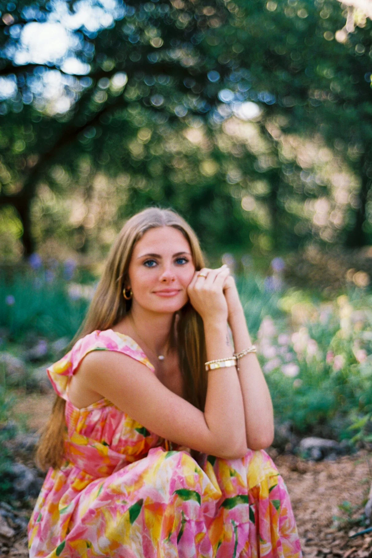A young woman with long, light brown hair and blue eyes, wearing a colorful, floral dress, sitting outdoors in a natural setting with trees and greenery in the background, with a relaxed expression.