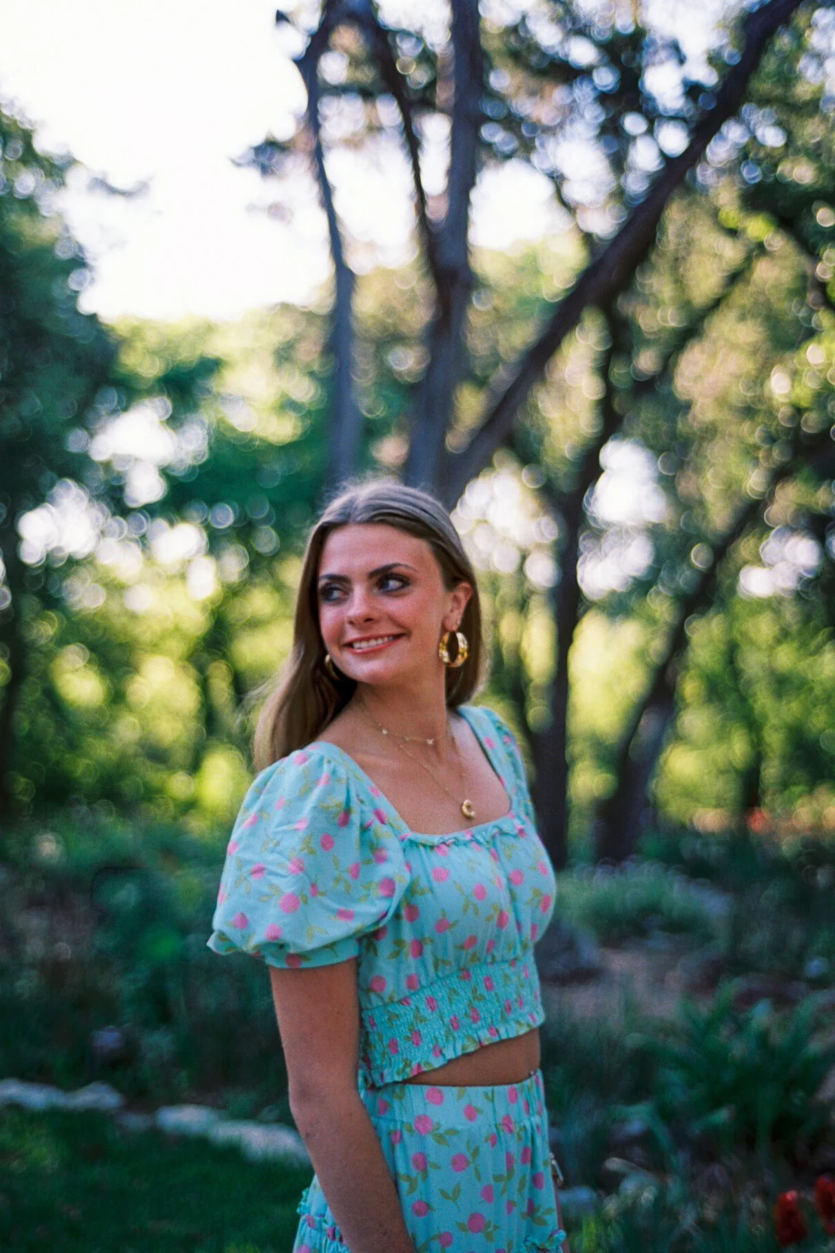 A young woman standing outdoors in a garden with trees in the background. She has long light brown hair and is smiling while looking to her left. She is wearing a light blue floral dress with puffed sleeves and layered gold jewelry.