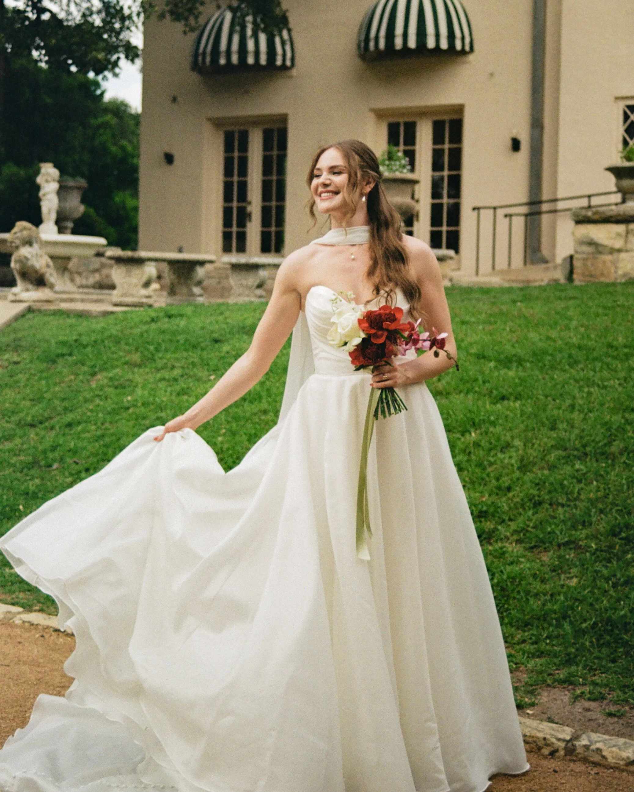 A smiling woman in a white wedding dress holding a bouquet of red and white flowers outdoors in front of a beige building with large windows and striped awnings.