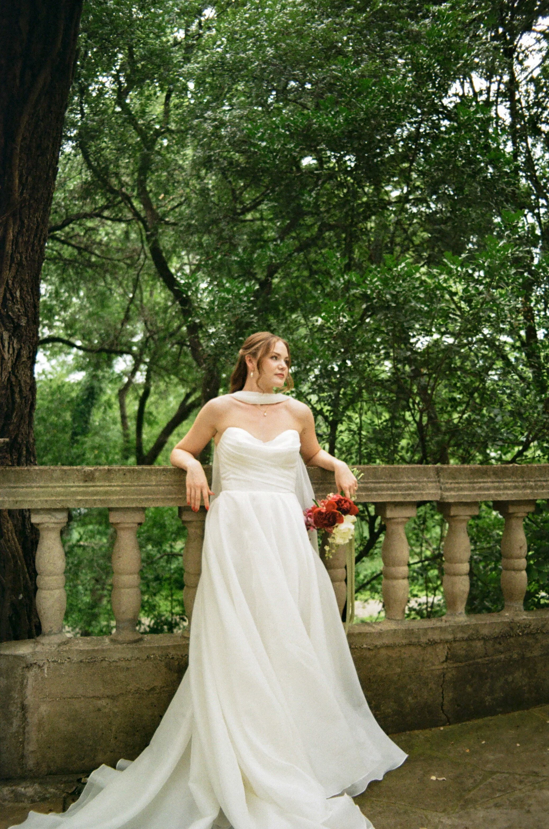 A bride in a white wedding gown holding a bouquet of flowers, standing on a stone staircase outdoors, surrounded by lush green trees.