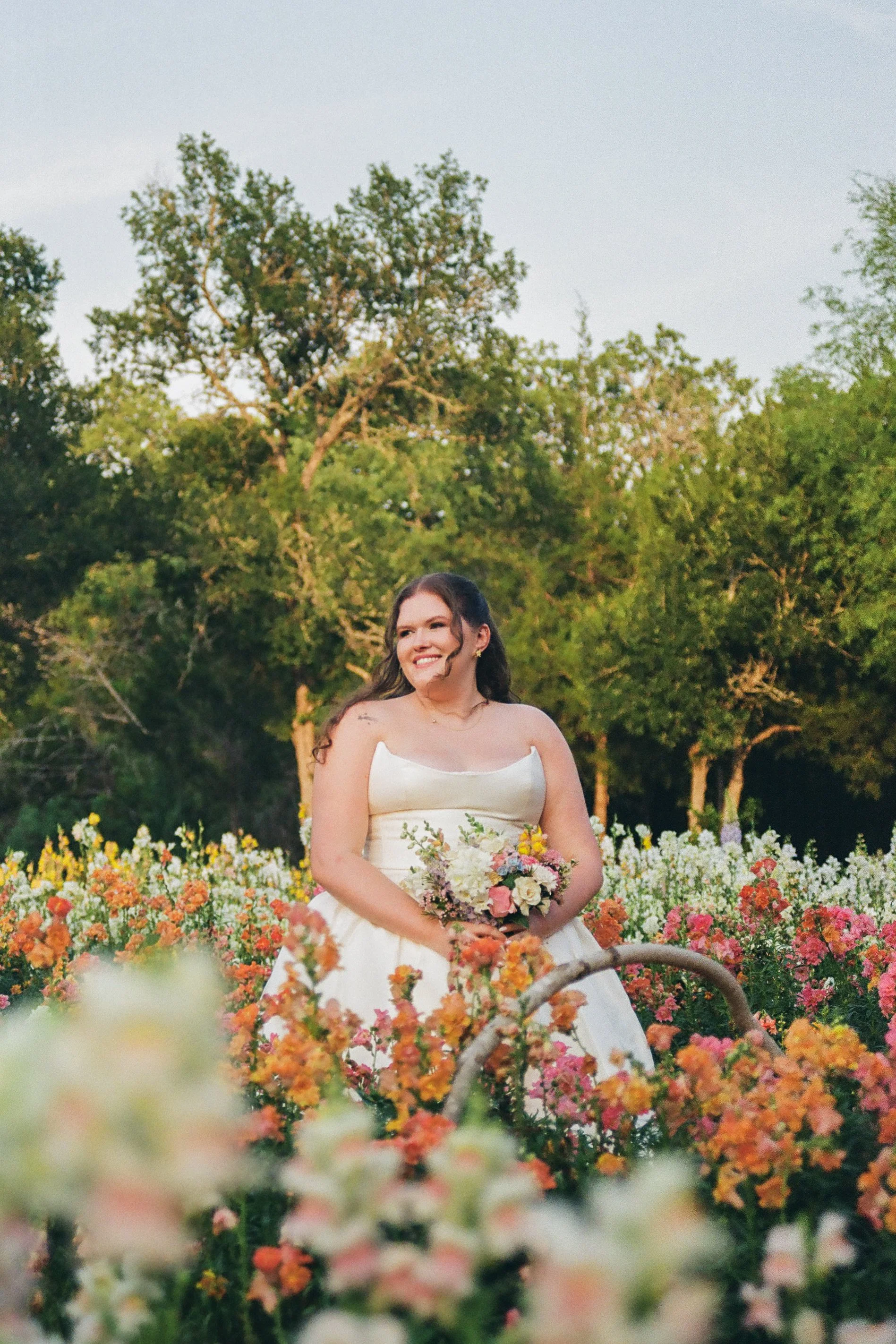 A bride in a white wedding gown holding a colorful bouquet, standing in a garden filled with orange, pink, and white flowers, with lush green trees in the background during sunset.