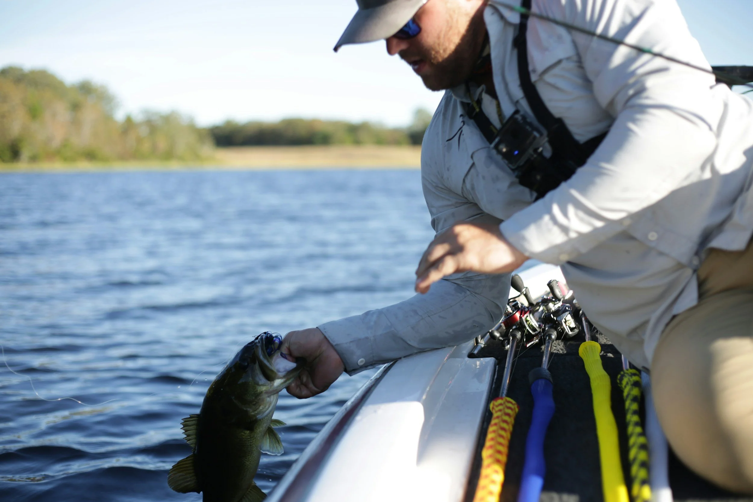 Hombre en ropa de pesca en kayak, sosteniendo un pez que acaba de atrapar en un cuerpo de agua, con fondo de árboles y cielo claro.