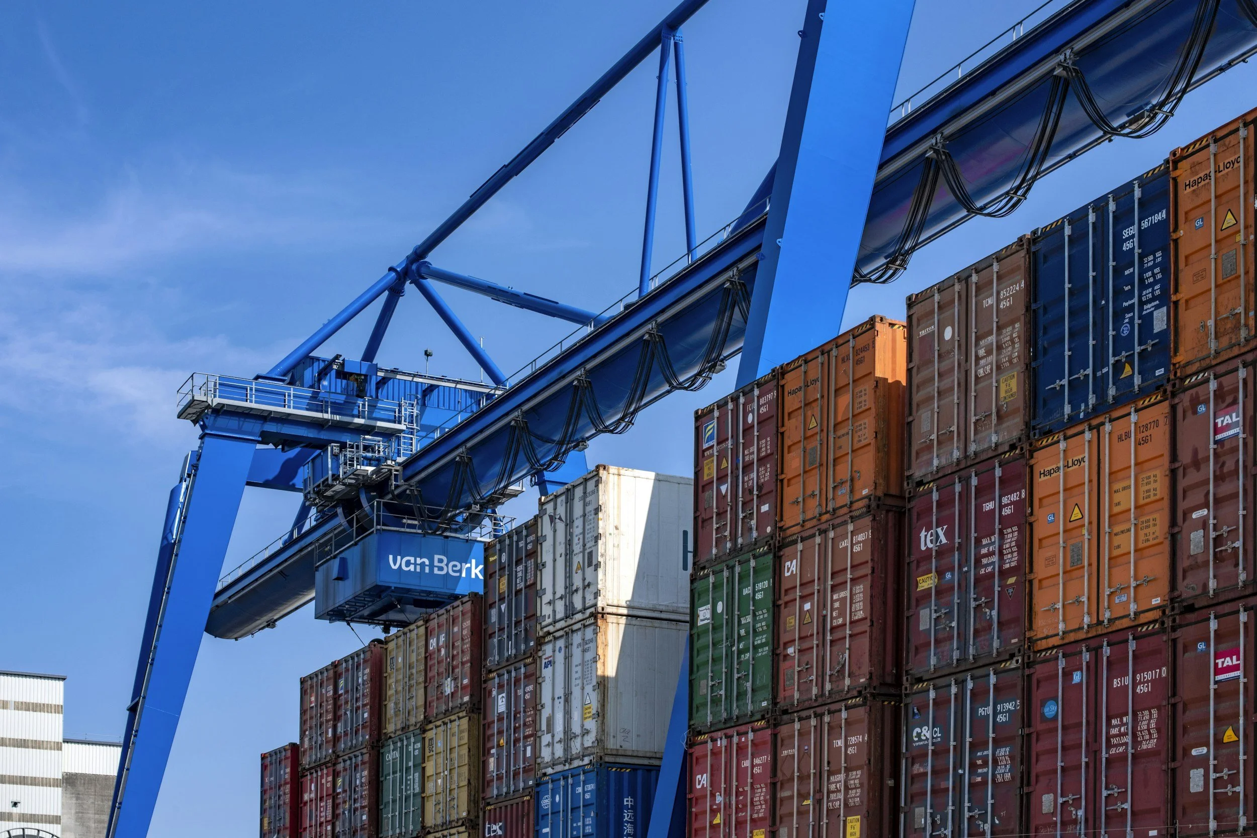 Un muelle de carga con contenedores de diferentes colores apilados y una grúa de color azul que los transporta bajo un cielo despejado.
