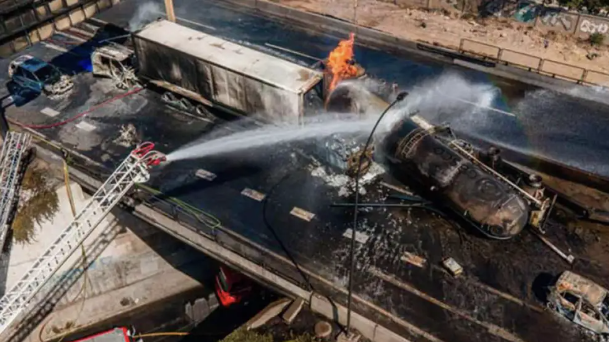 Incendio en un puente con vehículos accidentados, un camión ardiendo y un coche destruido, bomberos combaten las llamas.