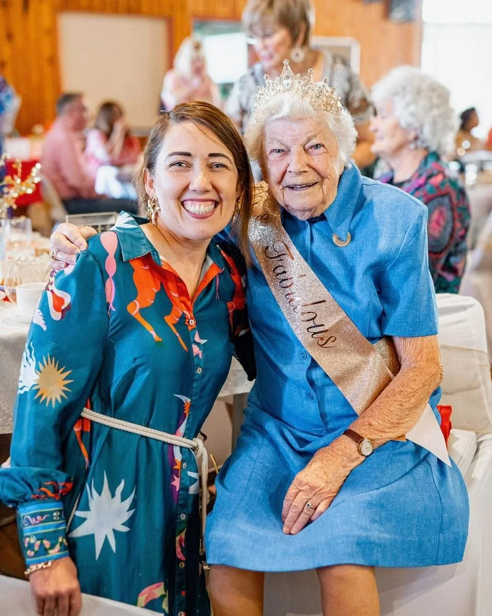 A woman wearing a blue dress with a sash that says "Grandma" is sitting and smiling, wearing a crown, at a celebration with many people in the background, some sitting at tables and others standing.